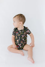 A baby sits on a white surface wearing the Jammie Session Mystic Mushrooms Short Sleeve Bodysuit, featuring a colorful mushroom and plant pattern, looking to the side.