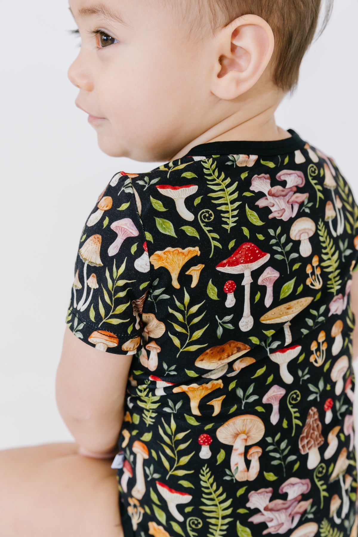 A young child wears the Jammie Session Mystic Mushrooms Short Sleeve Bodysuit featuring a colorful mushroom and fern design, sitting sideways against a plain background.