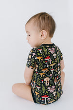 A toddler sits sideways on the floor in the Jammie Session Mystic Mushrooms Short Sleeve Bodysuit, featuring a vibrant mushroom and foliage pattern on black.
