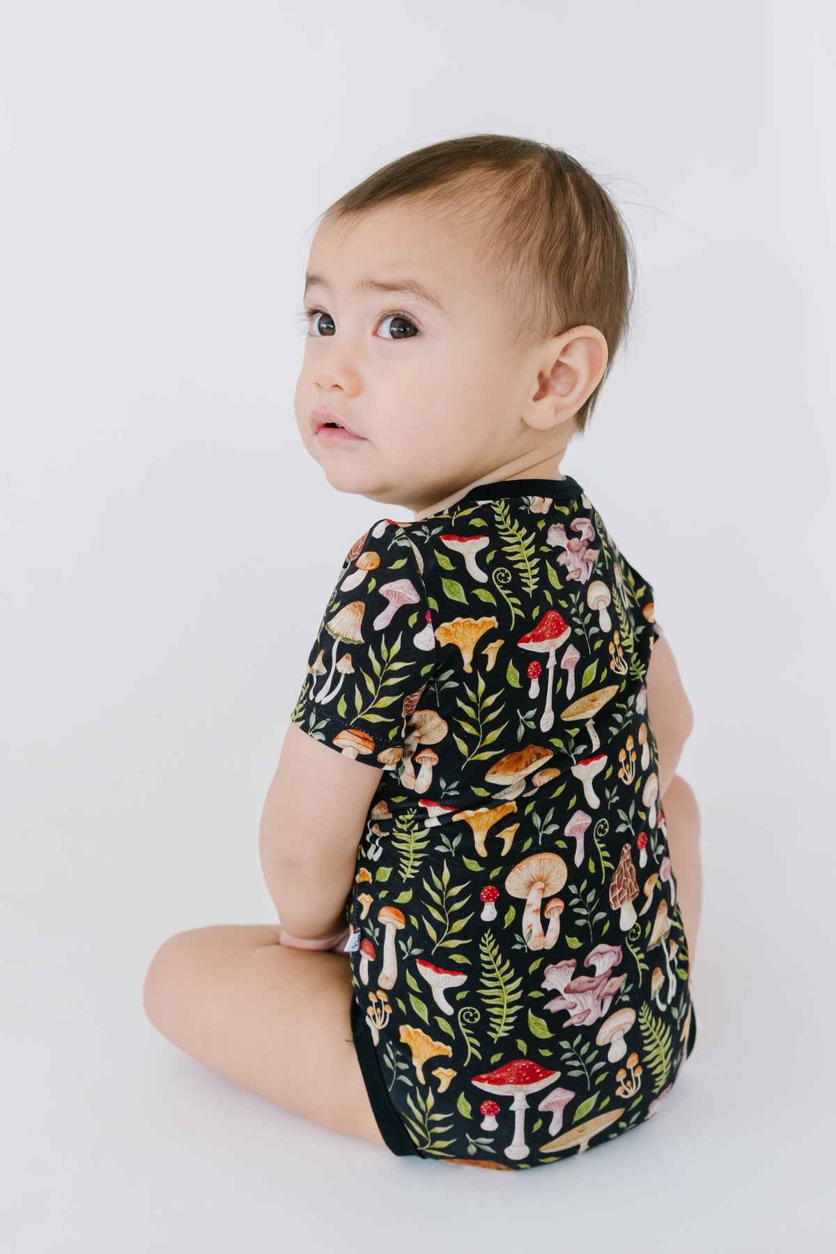 A baby wears the Jammie Session Mystic Mushrooms Short Sleeve Bodysuit, featuring colorful mushroom and leaf patterns, while sitting on a white background and looking over their shoulder.