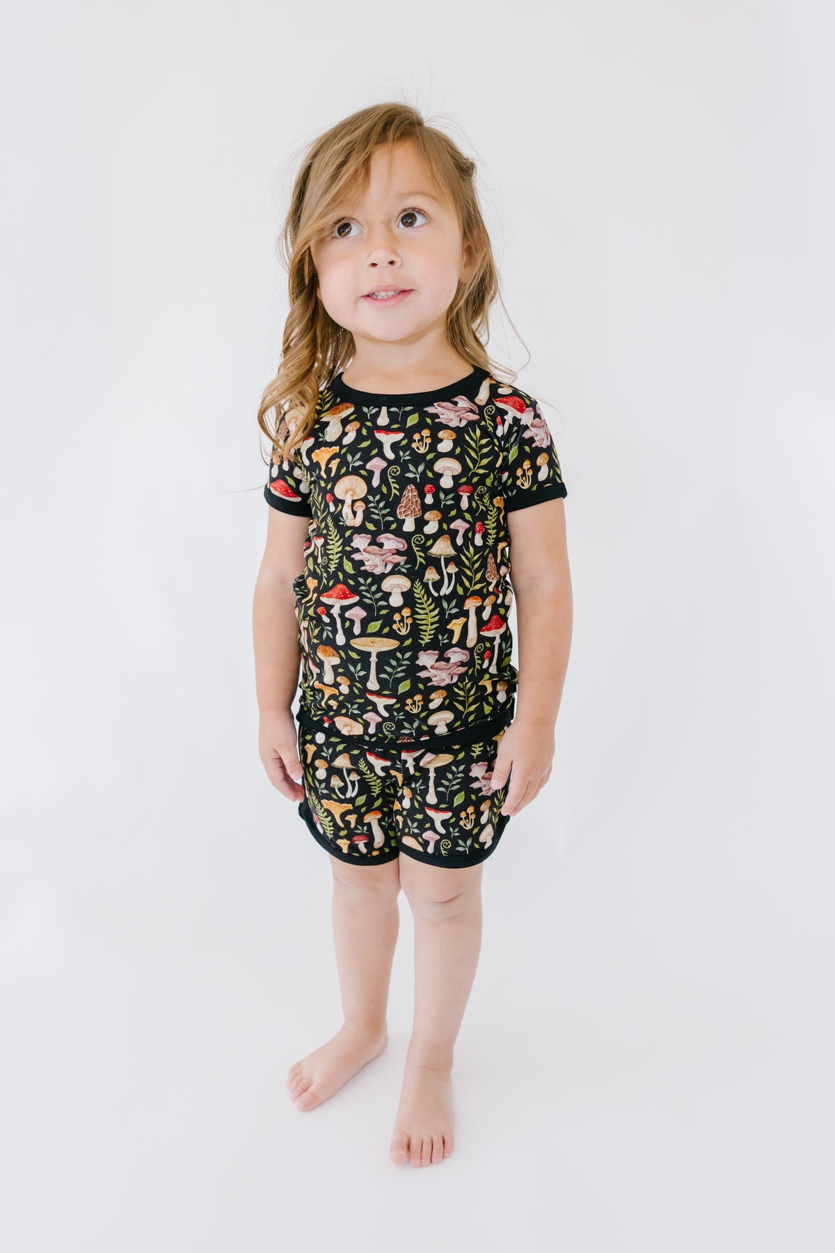 A young child stands against a white background, wearing the Jammie Session Mystic Mushrooms Short Pajama Set—soft, breathable kids pajamas in black with a vibrant mushroom print for all-day comfort.