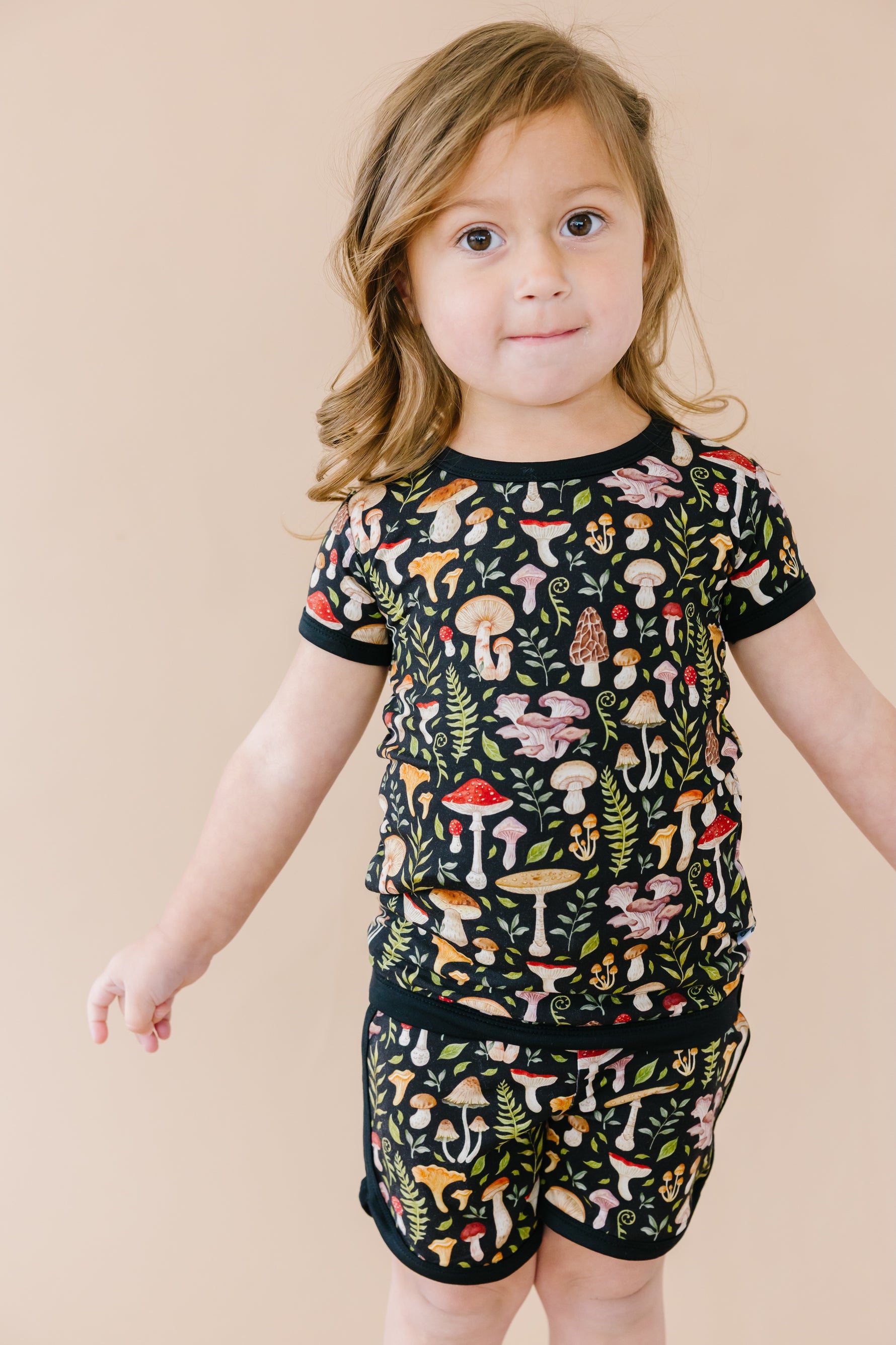 A young child wears the Jammie Session Mystic Mushrooms Short Pajama Set, featuring colorful mushroom and plant patterns, standing against a beige background.