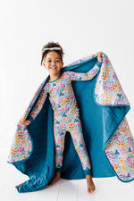 A young child smiles in matching floral pajamas and headband, holding open Jammie Session’s Whispering Wildflowers Toddler Quilted Blanket—a cozy, machine-washable 2.5 Tog bamboo quilt with a floral interior, perfect for bedtime.