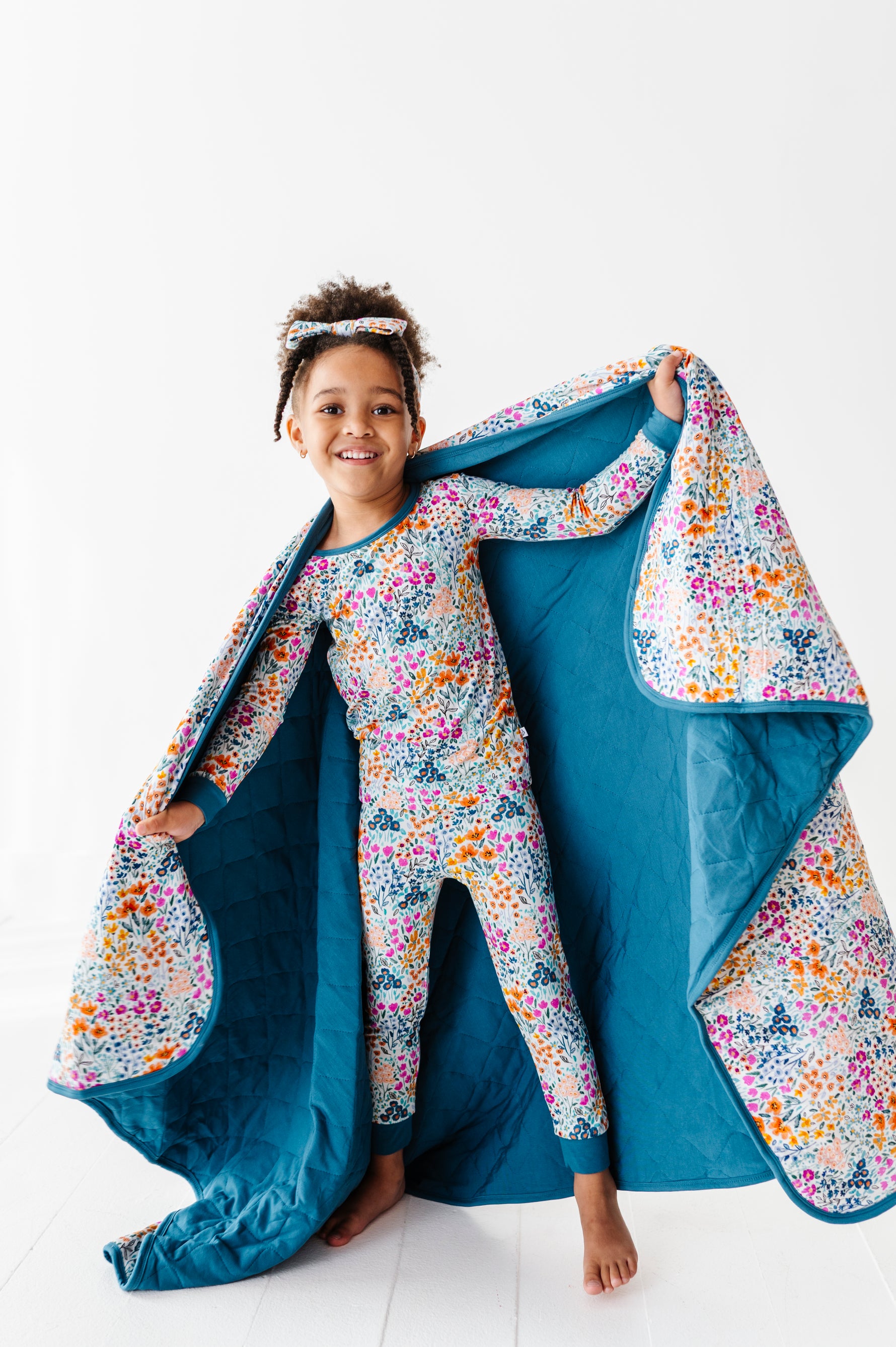 A young child smiles in matching floral pajamas and headband, holding open Jammie Session’s Whispering Wildflowers Toddler Quilted Blanket—a cozy, machine-washable 2.5 Tog bamboo quilt with a floral interior, perfect for bedtime.