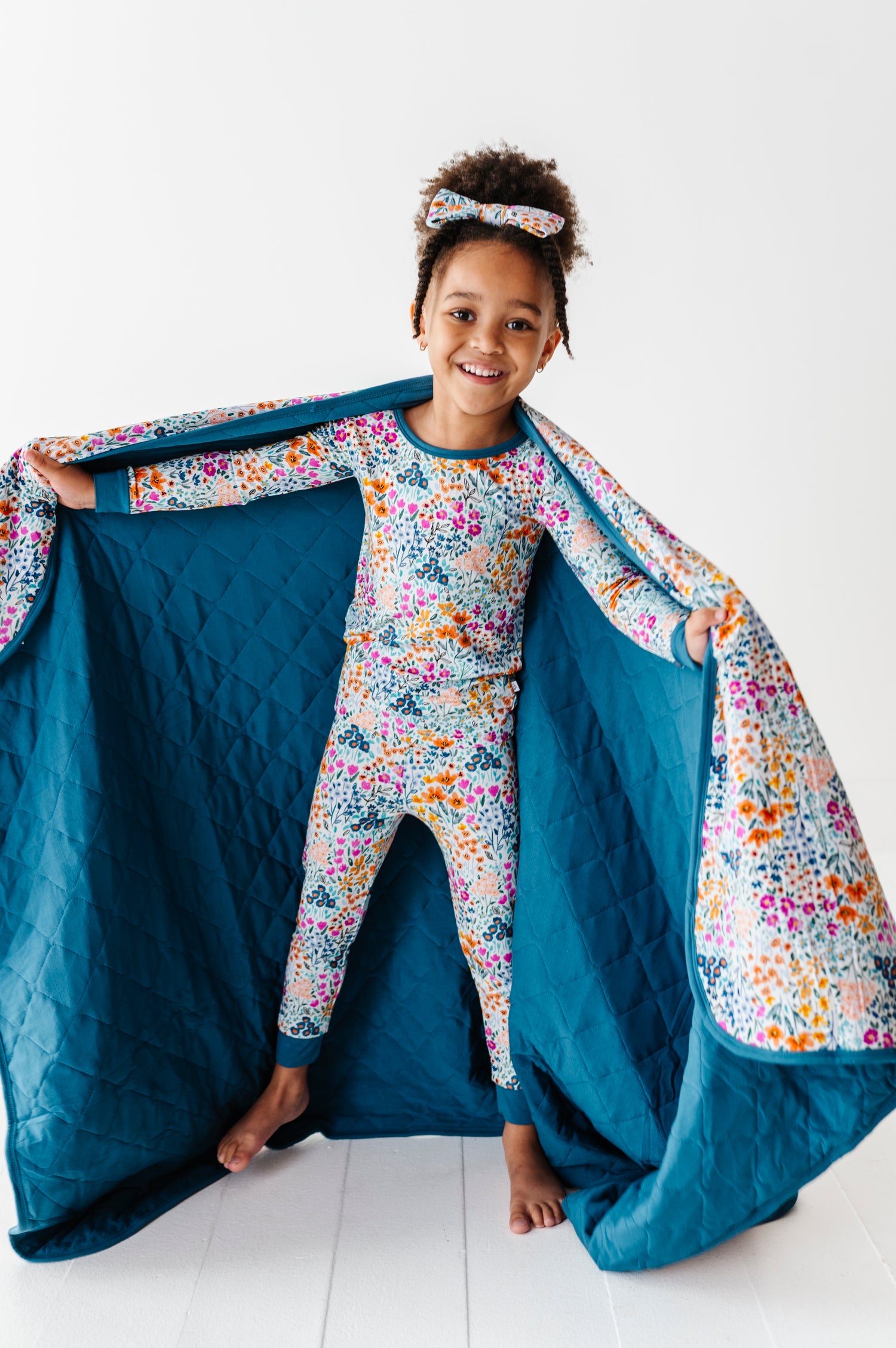 A young girl stands inside the open, machine-washable Whispering Wildflowers Toddler Quilted Blanket by Jammie Session, smiling at the camera in her colorful floral pajama set and matching headband.