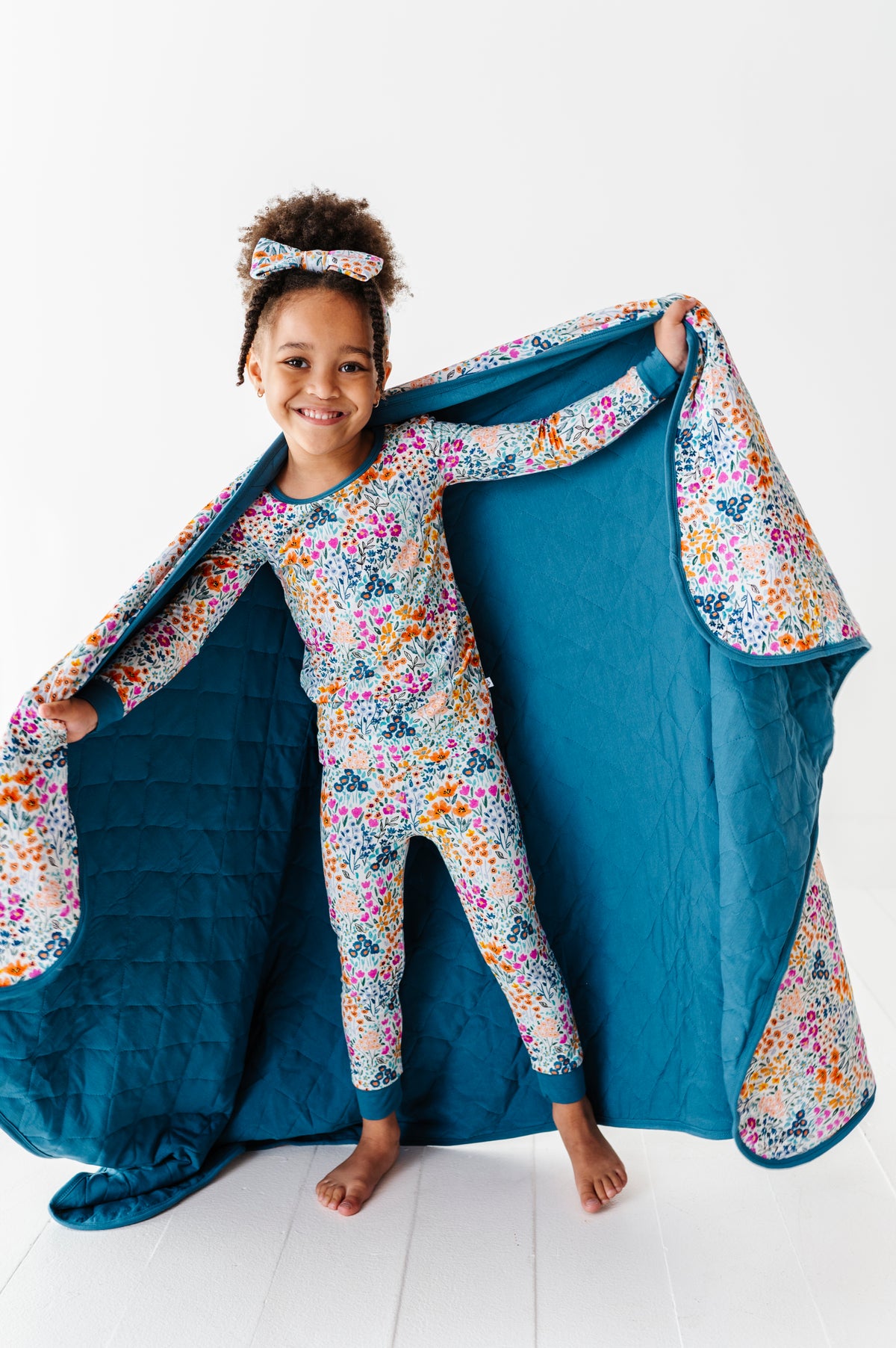 A smiling young girl in matching floral pajamas and headband holds open the Jammie Session Whispering Wildflowers Toddler Quilted Blanket, showing its soft, machine washable blue exterior and inner floral pattern against a white background.