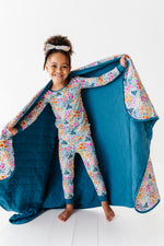 A smiling young girl in matching floral pajamas and headband holds open the Jammie Session Whispering Wildflowers Toddler Quilted Blanket, showing its soft, machine washable blue exterior and inner floral pattern against a white background.