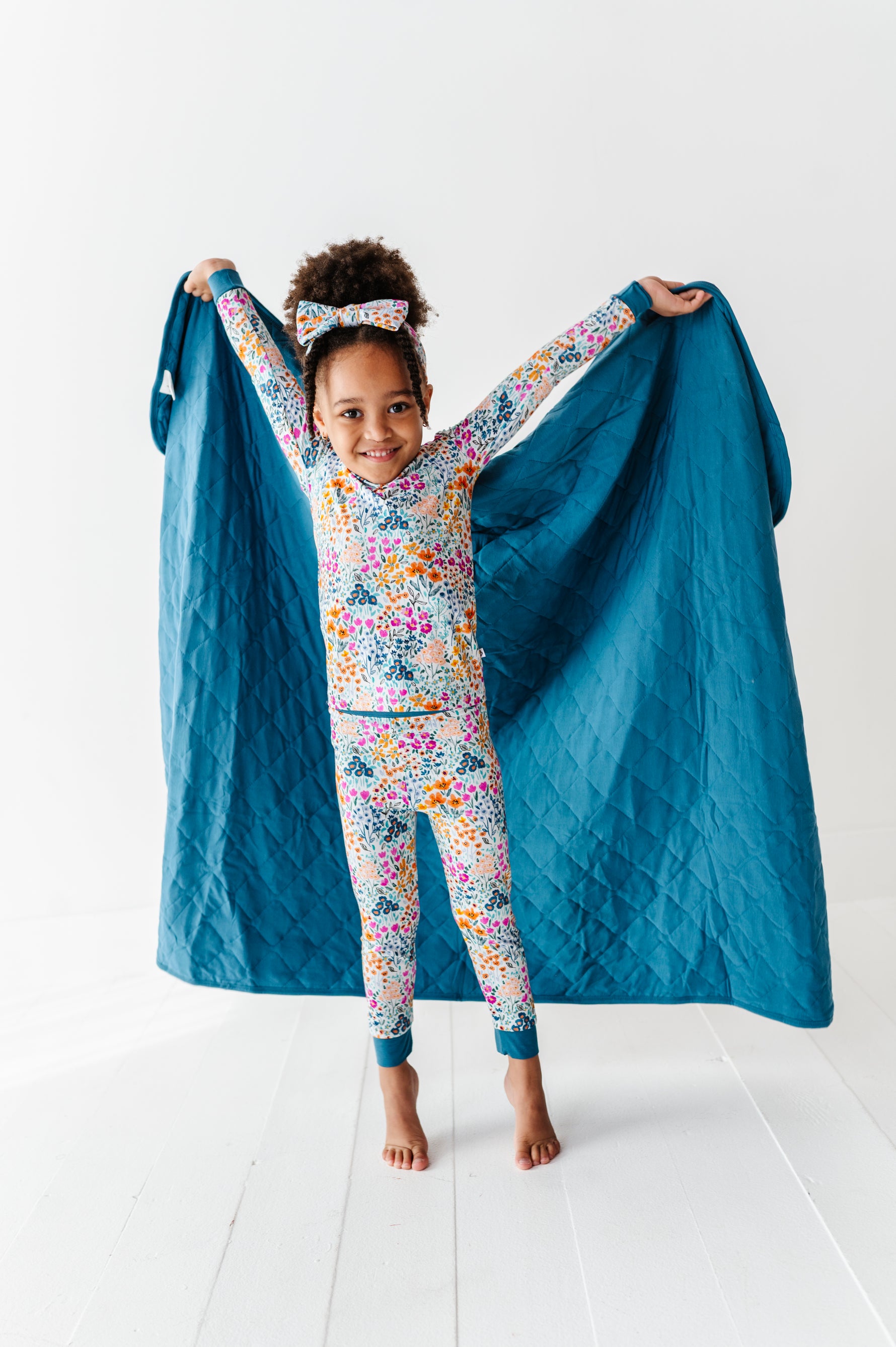 A young girl in colorful floral pajamas and a matching headband stands barefoot, holding up the Jammie Session Whispering Wildflowers Toddler Quilted Blanket—large, blue, bamboo, and machine washable—on a white floor.
