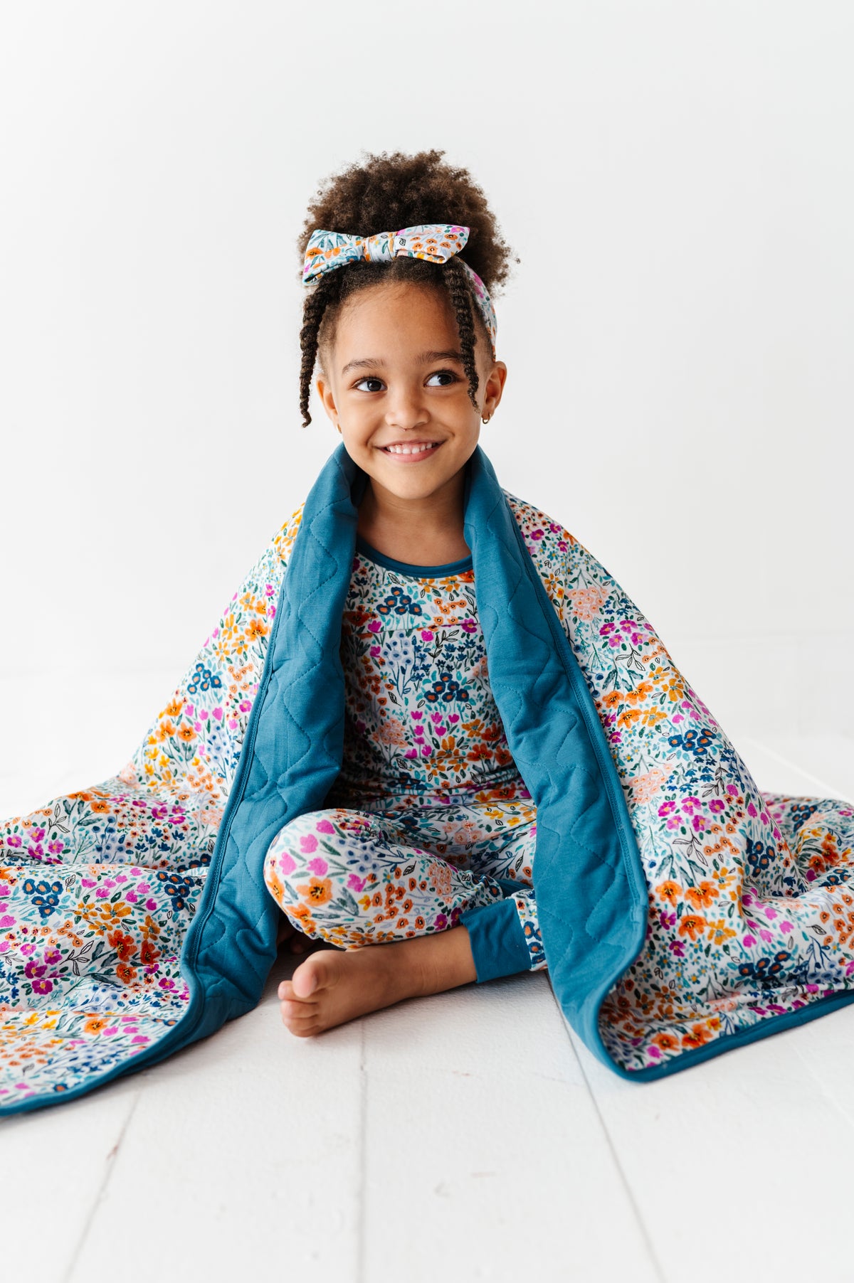 A young girl sits cross-legged on the floor in floral pajamas and a matching headband, wrapped in the Jammie Session Whispering Wildflowers Toddler Quilted Blanket with a blue border.
