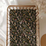 The Jammie Session Mystic Mushrooms Crib Sheet offers soft, hypoallergenic baby bedding with a colorful mushroom and botanical print, shown here in a wooden crib with a dark blanket and dried flowers in a softly lit room.
