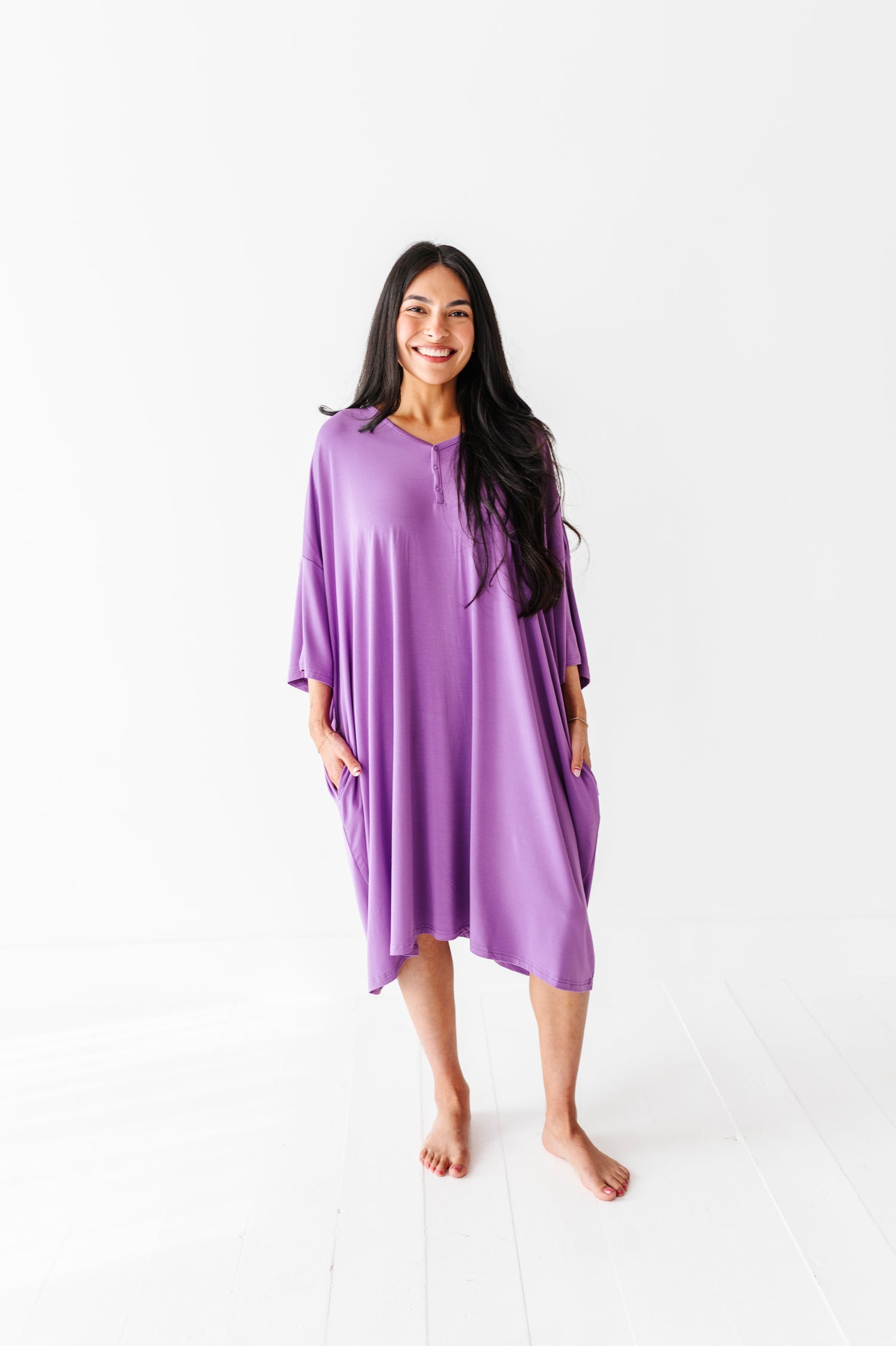 A woman with long dark hair stands barefoot, smiling, wearing the Jammie Session Block Party Solid Women's Muumuu Lounge Tee (ONE SIZE FITS XS - XXL, purple with pockets) against a white background.