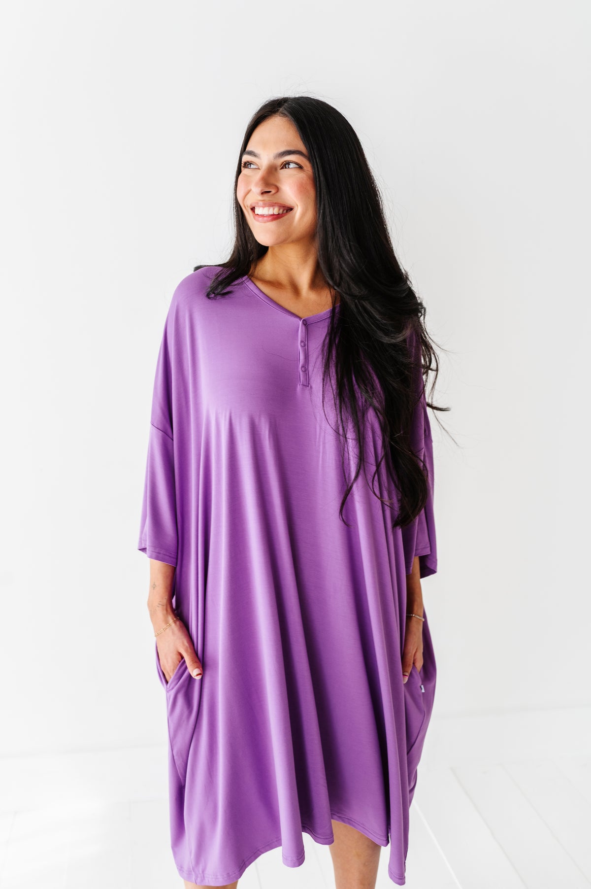 A woman wearing the Jammie Session Block Party Solid Women's Muumuu Lounge Tee (ONE SIZE FITS XS - XXL) in purple stands against a white background, smiling to the side with her hands in her pockets.