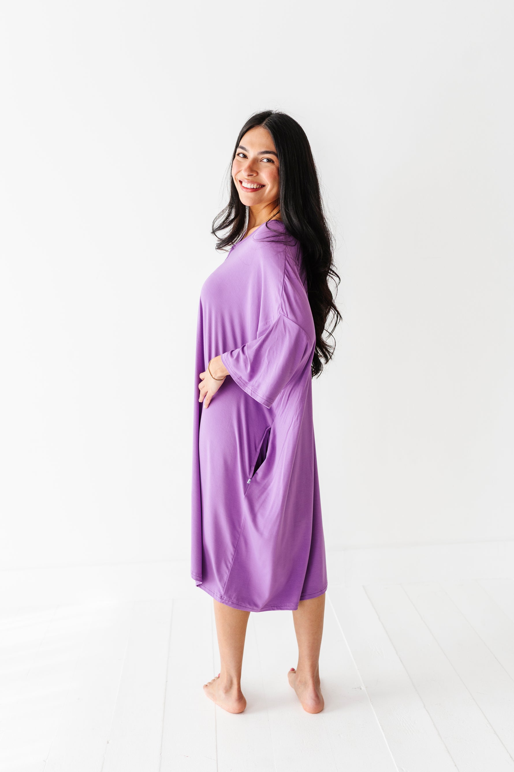 A woman with long dark hair smiles at the camera, barefoot on a white floor, wearing the Jammie Session Block Party Solid Women's Muumuu Lounge Tee (ONE SIZE FITS XS - XXL), showcasing its relaxed and comfortable oversized fit.