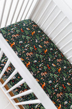 A white crib with a mattress covered by the Jammie Session Wood You Be My Friend Crib Sheet, featuring dark green, breathable baby bedding with a colorful forest animal and plant pattern.