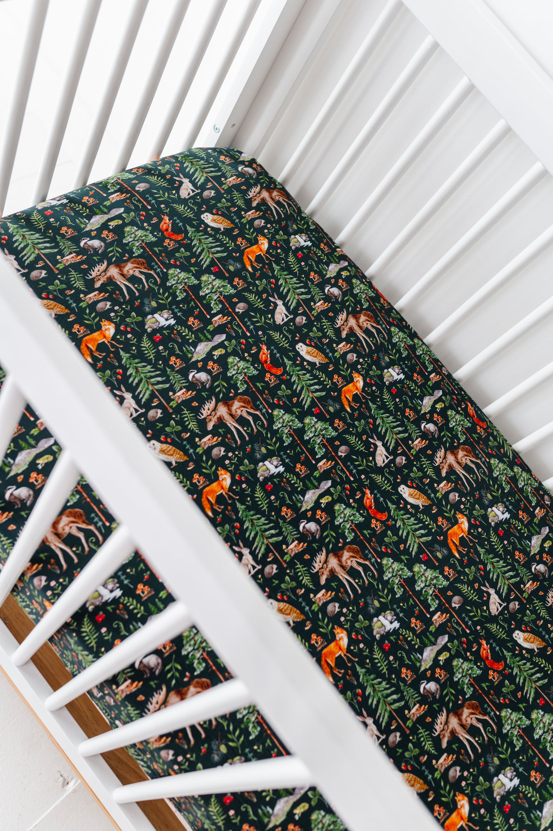 A white crib with a mattress covered by the Jammie Session Wood You Be My Friend Crib Sheet, featuring dark green, breathable baby bedding with a colorful forest animal and plant pattern.