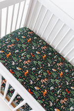 A white crib with the Jammie Session "Wood You Be My Friend" crib sheet, featuring breathable fabric and a fitted design decorated with colorful forest animals and leaves on a dark background.