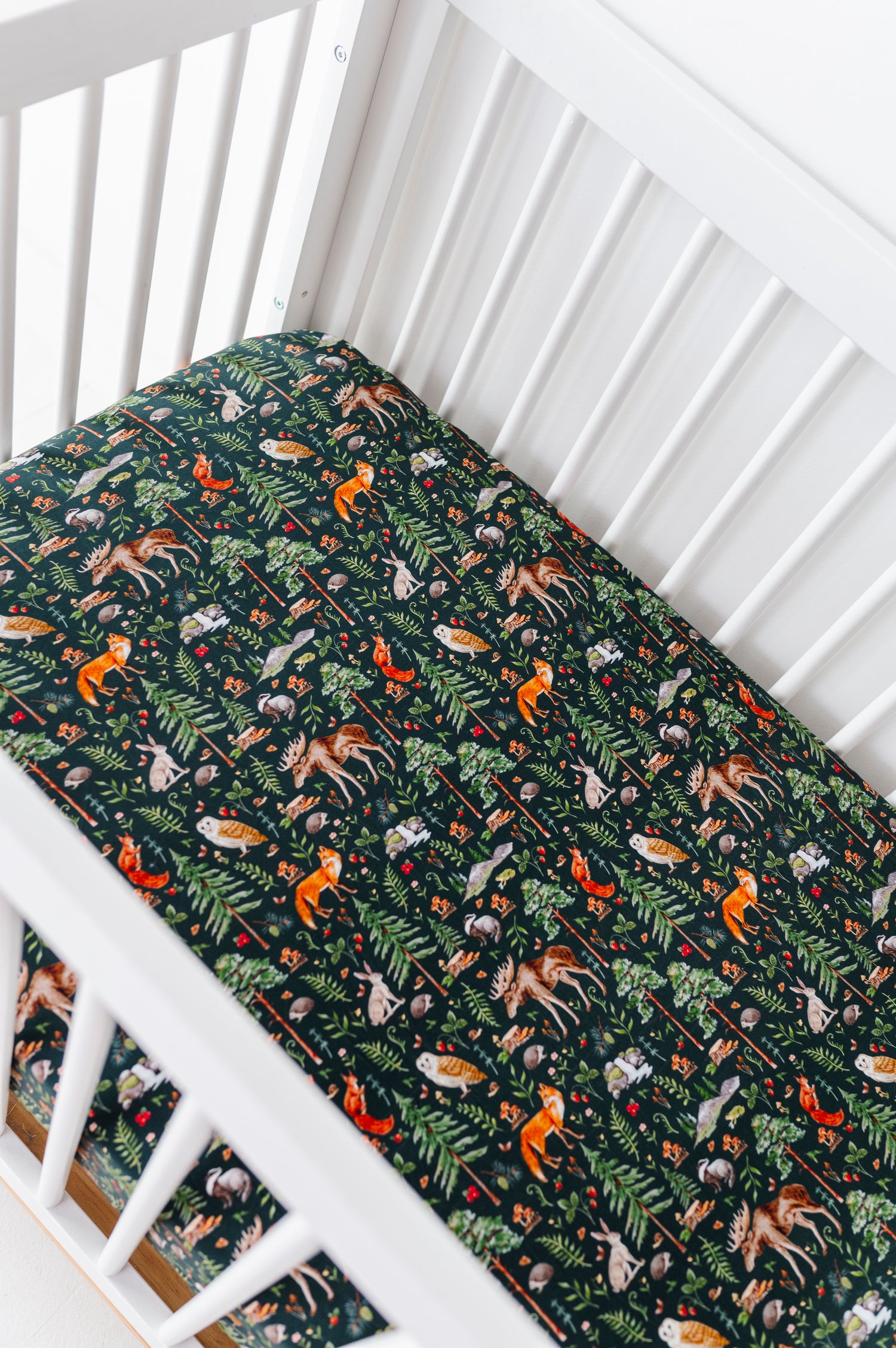 A white crib with the Jammie Session "Wood You Be My Friend" crib sheet, featuring breathable fabric and a fitted design decorated with colorful forest animals and leaves on a dark background.