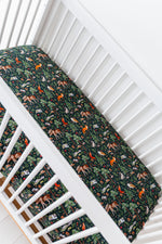 A white crib with a mattress dressed in the Jammie Session "Wood You Be My Friend Crib Sheet," featuring dark blue, breathable fabric adorned with colorful woodland animals and foliage.