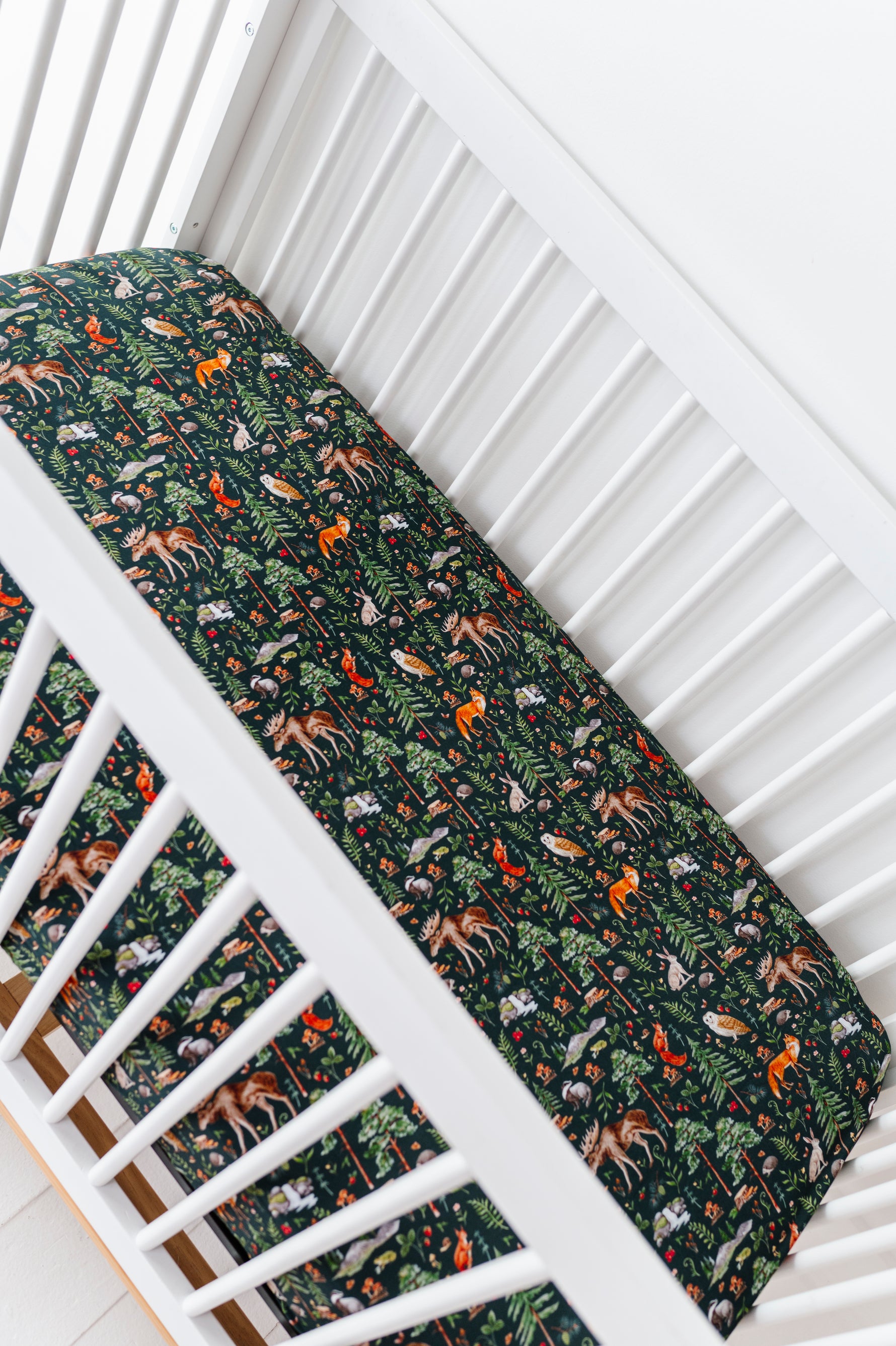A white crib with a mattress dressed in the Jammie Session "Wood You Be My Friend Crib Sheet," featuring dark blue, breathable fabric adorned with colorful woodland animals and foliage.