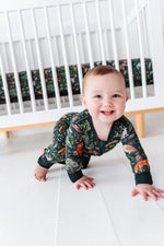 A smiling baby crawls on a white floor in front of a crib with the Jammie Session Wood You Be My Friend Crib Sheet—breathable, hypoallergenic, and adorned with matching animal print.