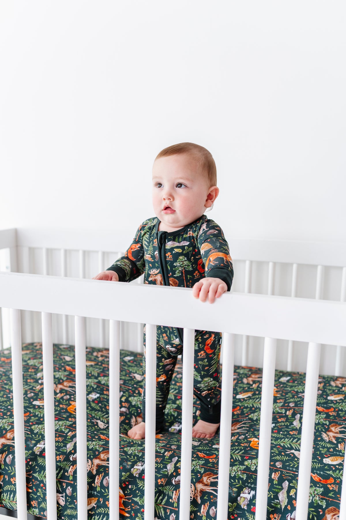 A baby stands in a white crib, wearing a dark animal-print onesie that matches the Jammie Session "Wood You Be My Friend" Crib Sheet—perfect breathable bedding for your jungle-themed nursery.
