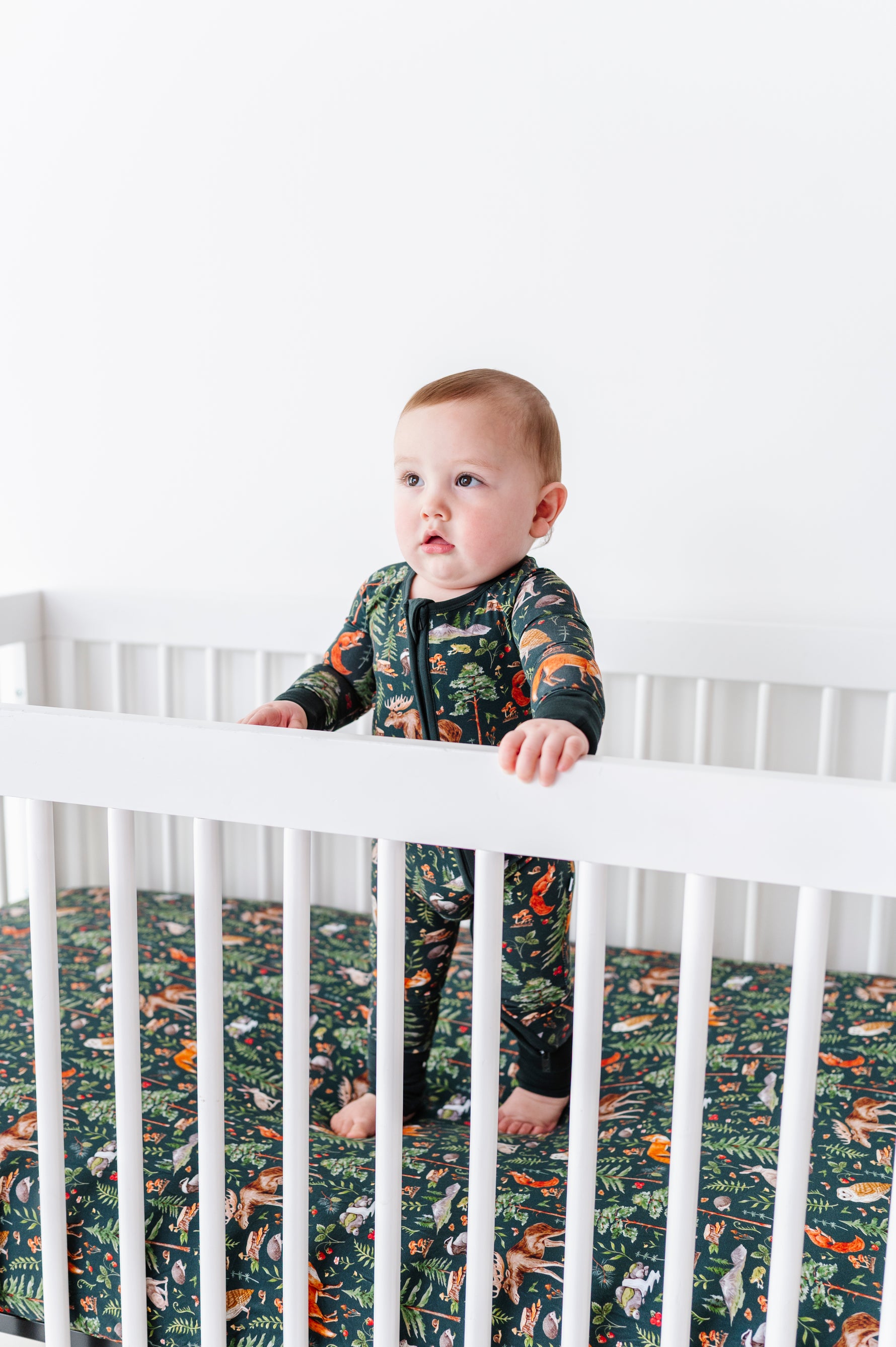 A baby stands in a white crib, wearing a dark animal-print onesie that matches the Jammie Session "Wood You Be My Friend" Crib Sheet—perfect breathable bedding for your jungle-themed nursery.
