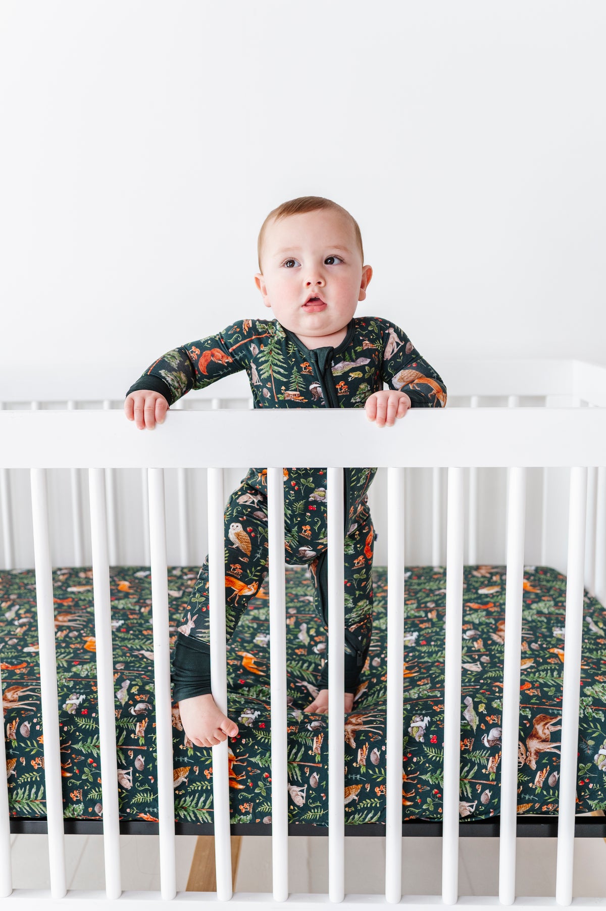 A baby in a patterned onesie stands inside a white crib, holding the rails. The mattress features the Jammie Session Wood You Be My Friend Crib Sheet for hypoallergenic, jungle-themed comfort and safety.