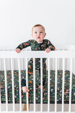A baby in a patterned onesie stands inside a white crib, holding the rails. The mattress features the Jammie Session Wood You Be My Friend Crib Sheet for hypoallergenic, jungle-themed comfort and safety.