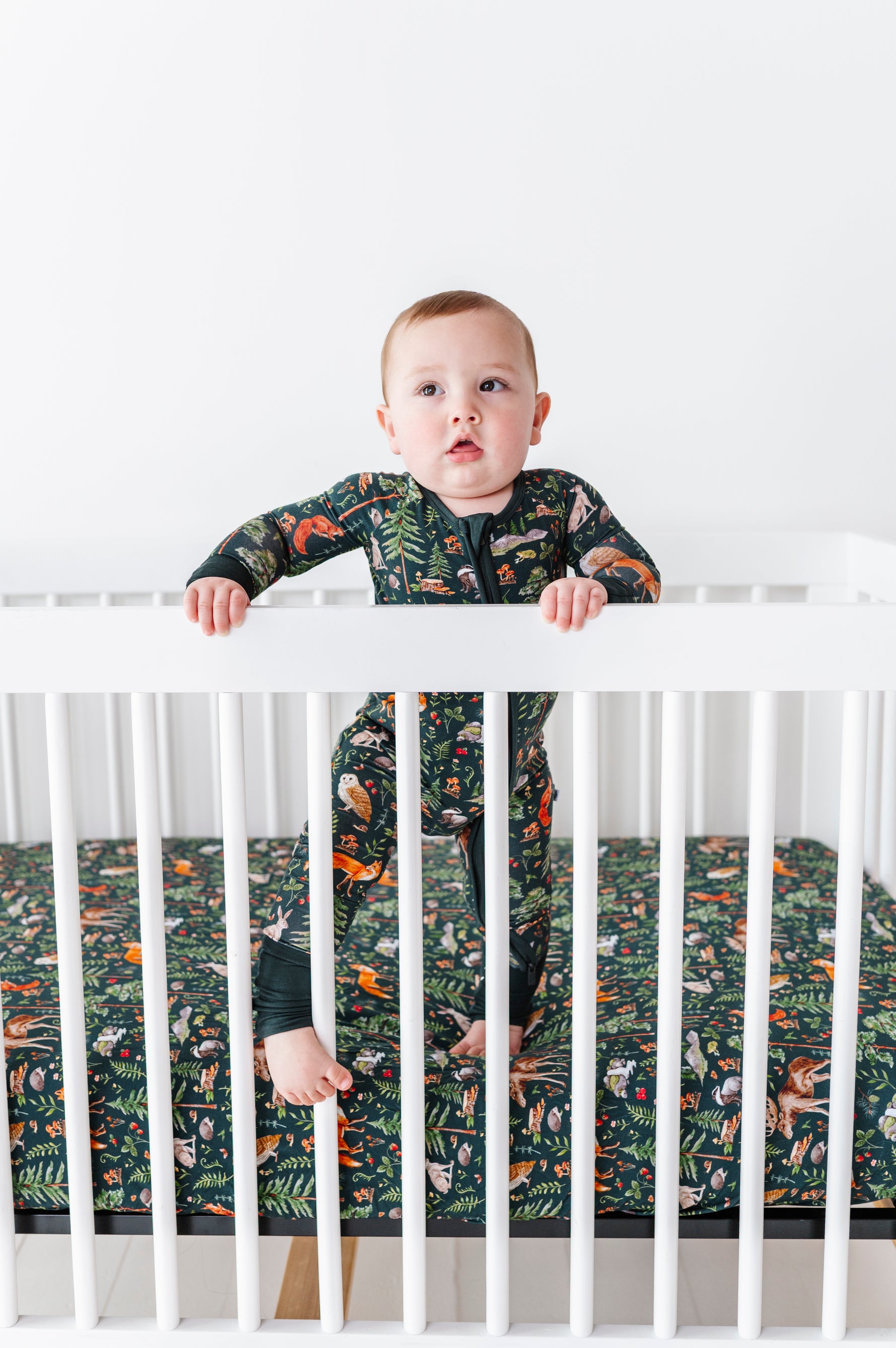 A baby in a patterned onesie stands inside a white crib, holding the rails. The mattress features the Jammie Session Wood You Be My Friend Crib Sheet for hypoallergenic, jungle-themed comfort and safety.