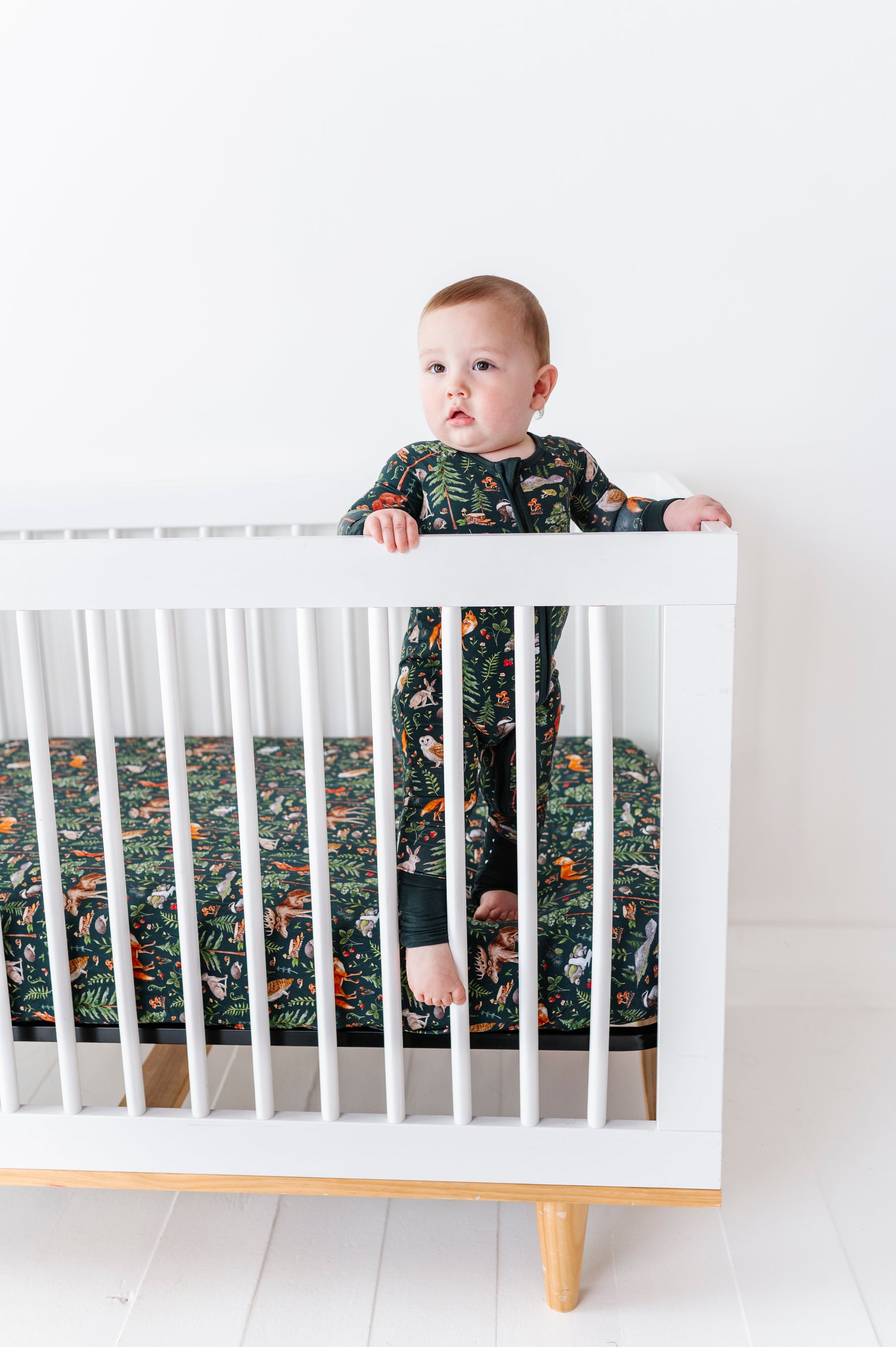 A baby in dark patterned pajamas stands by a white crib, matching the Jammie Session Wood You Be My Friend Crib Sheet, in a bright, minimal room with breathable bedding.
