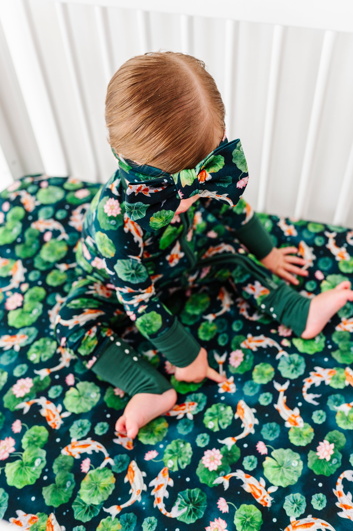A toddler in green floral pajamas with a matching headband sits on a Jammie Session Bundle of Koi Crib Sheet, featuring an ultra-soft bamboo fabric with lotus and koi fish patterns.
