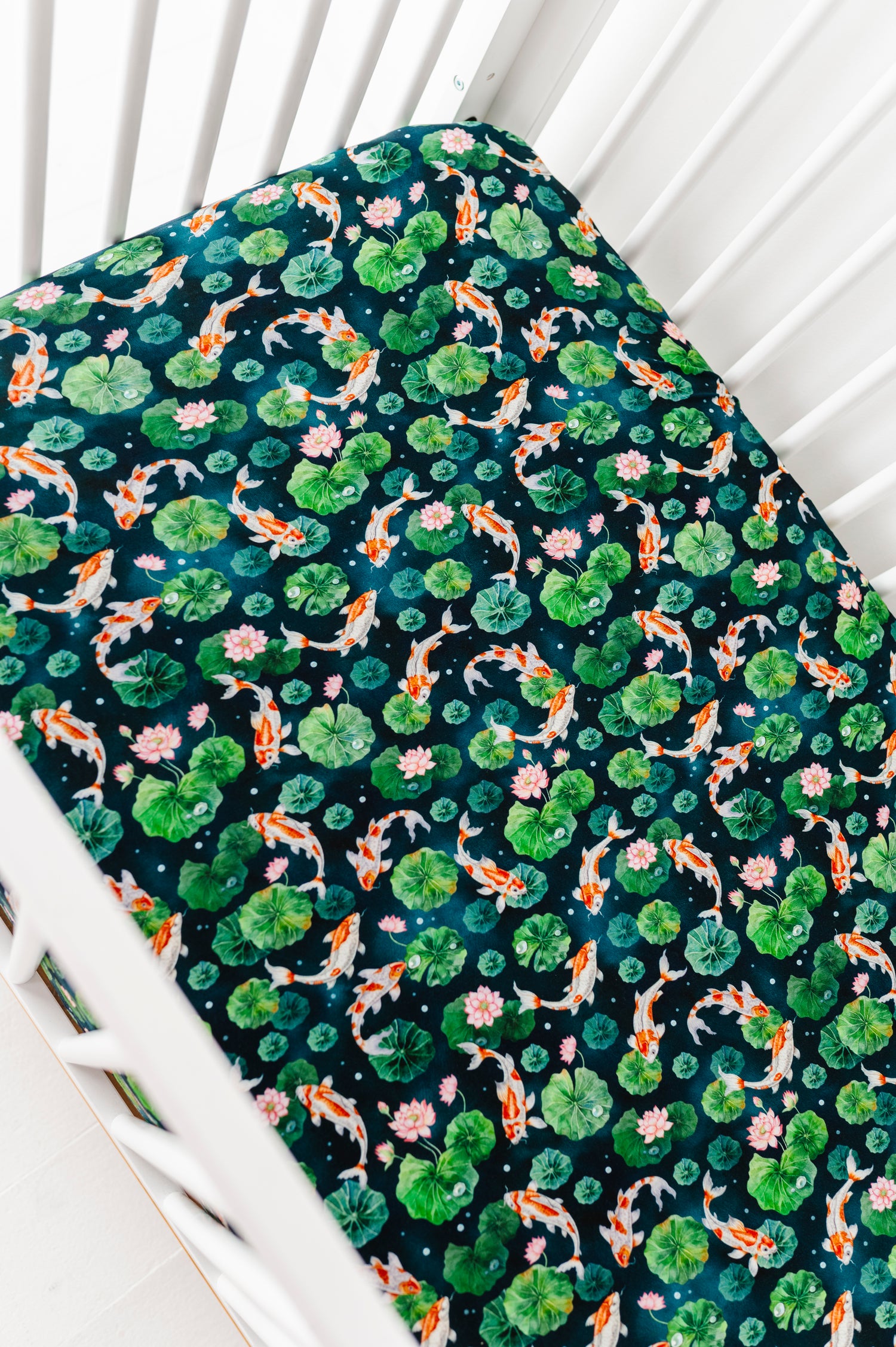 The Jammie Session Bundle of Koi Crib Sheet covers the mattress on this white crib, featuring an ultra soft, hypoallergenic fabric with koi fish, lily pads, and pink flowers on a dark background.