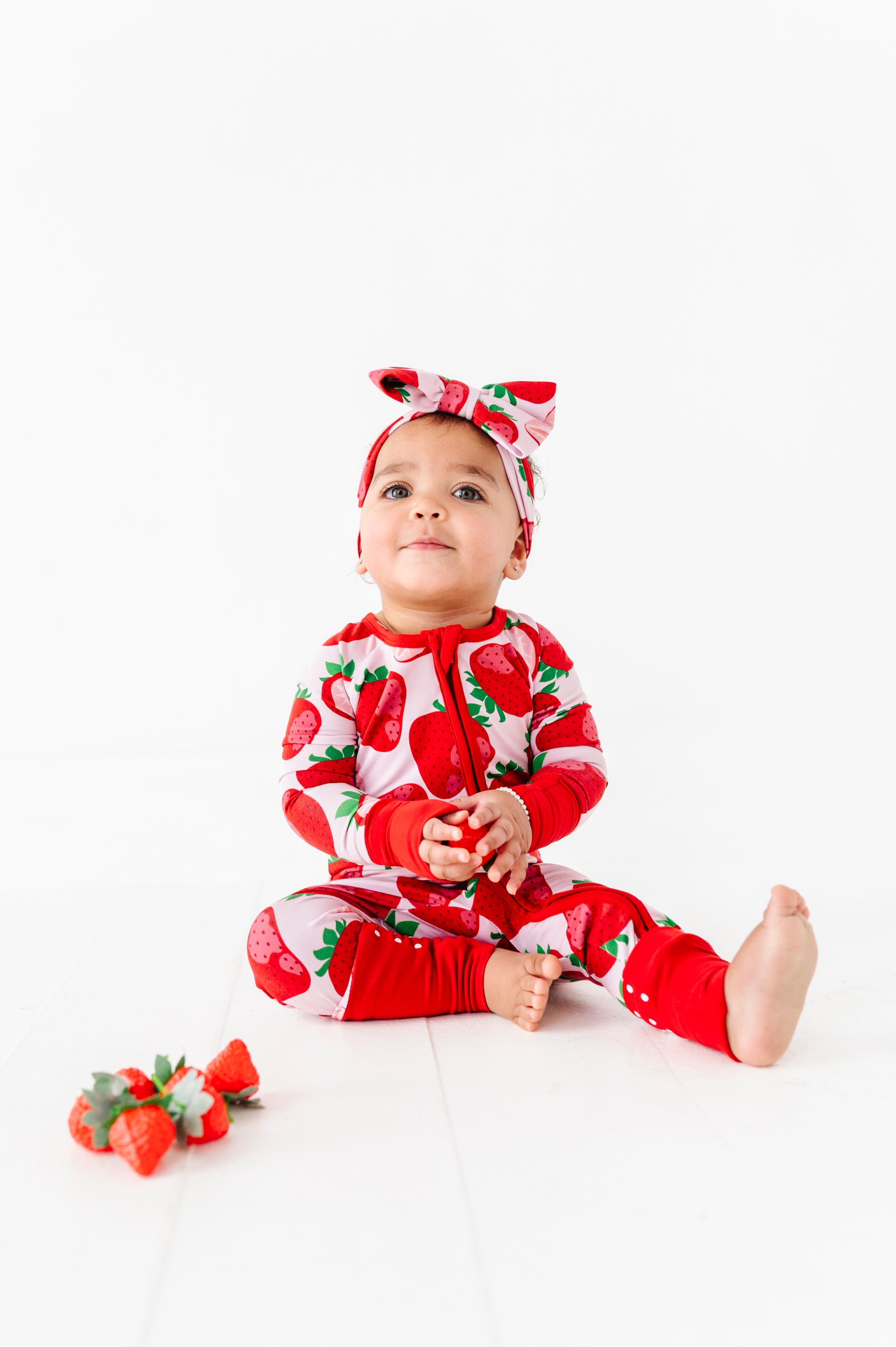 A baby in the Jammie Session Berry Sweet (Strawberry) Romper with matching headband sits on a white floor, hands clasped, near a small pile of strawberries.