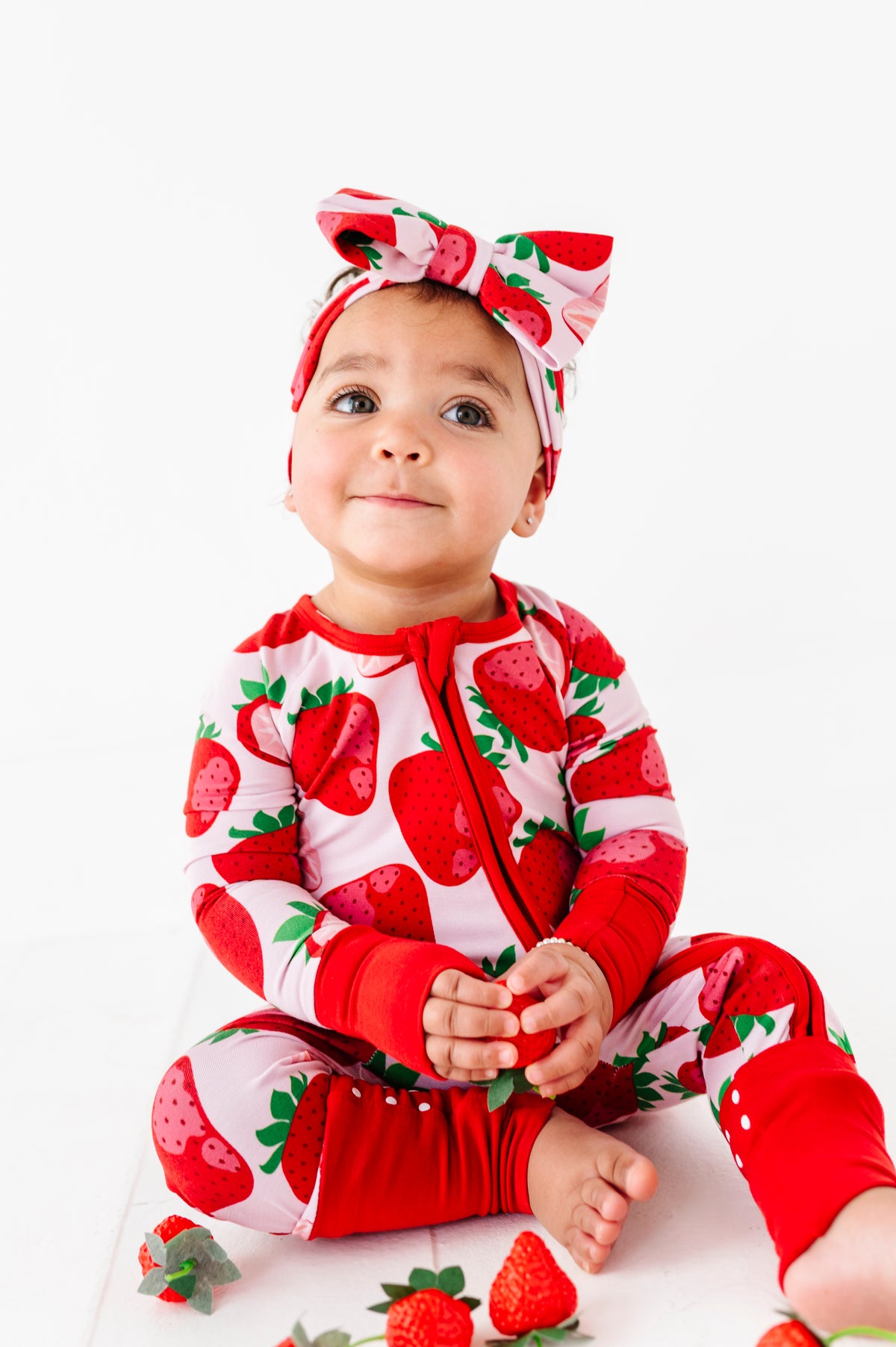 A baby wears the Jammie Session Berry Sweet (Strawberry) Romper and matching headband, sitting on the floor holding a strawberry, with more strawberries scattered around them on a white background.