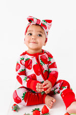 A baby wears the Jammie Session Berry Sweet (Strawberry) Romper and matching headband, sitting on the floor holding a strawberry, with more strawberries scattered around them on a white background.