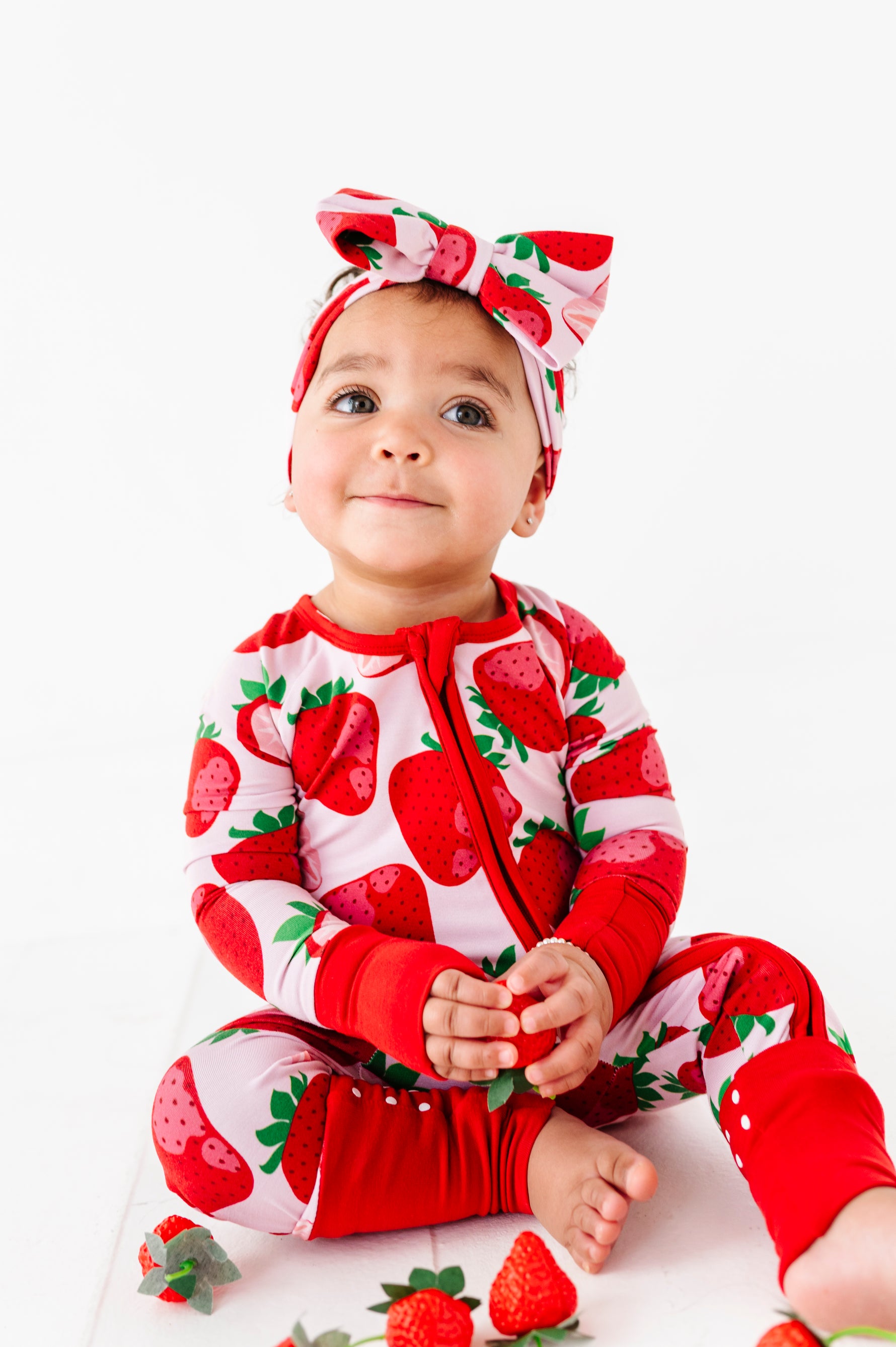A baby wears the Jammie Session Berry Sweet (Strawberry) Romper and matching headband, sitting on the floor holding a strawberry, with more strawberries scattered around them on a white background.