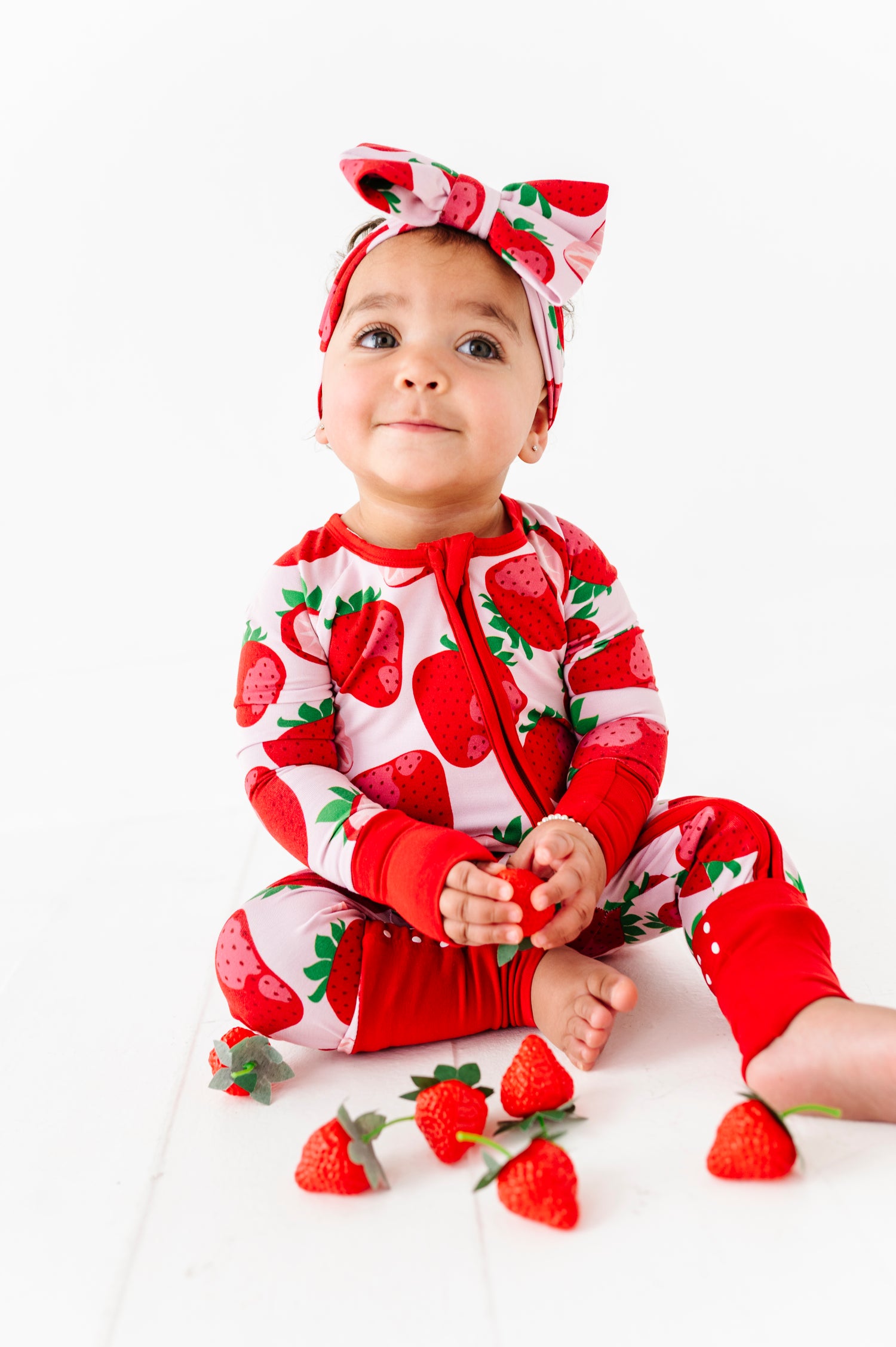 A baby in the Berry Sweet (Strawberry) Romper and matching headband by Jammie Session sits on the floor, holding strawberries, with more strawberries placed in front.