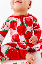 A baby in a Berry Sweet (Strawberry) Romper by Jammie Session sits on a white surface, smiling with their mouth closed.