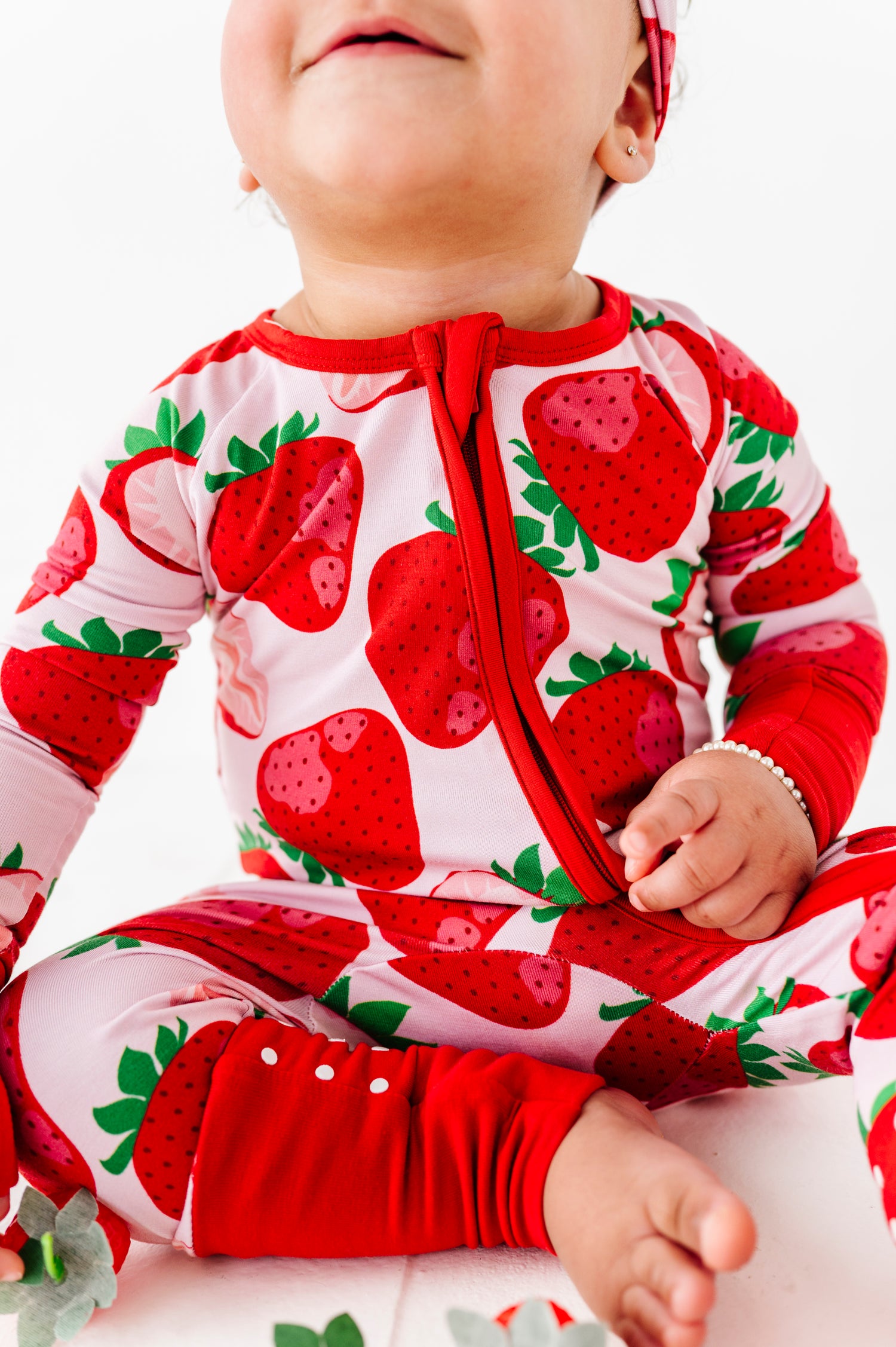 A baby in a Berry Sweet (Strawberry) Romper by Jammie Session sits on a white surface, smiling with their mouth closed.