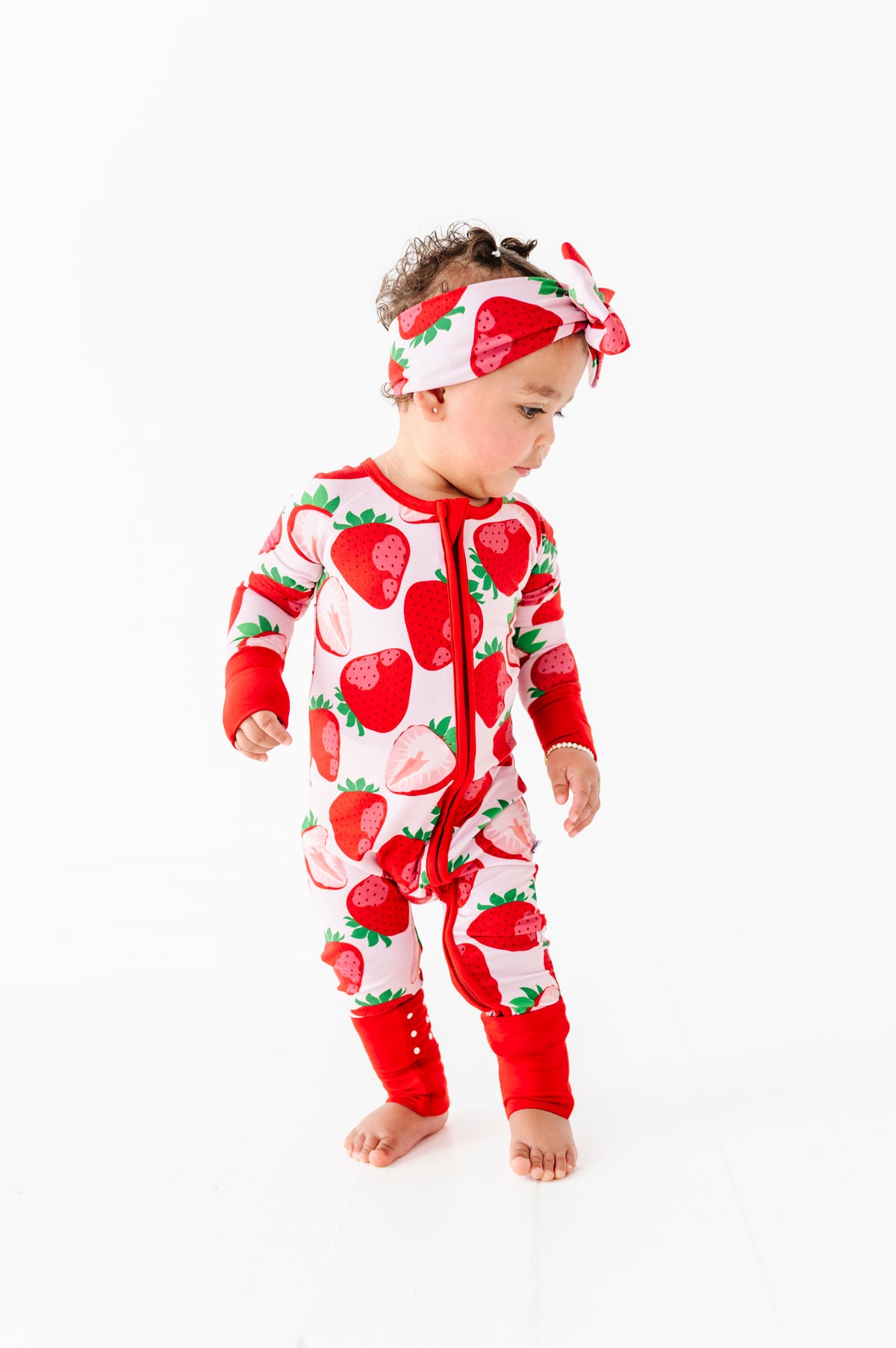 A toddler in a Jammie Session Berry Sweet (Strawberry) Romper with a matching headband stands against a plain white background.