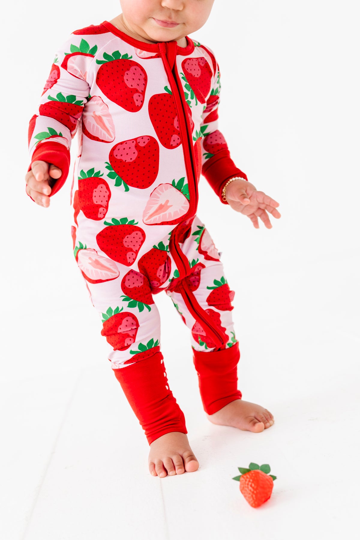 A baby in a Jammie Session Berry Sweet (Strawberry) Romper with long sleeves stands barefoot on a white surface beside a toy strawberry.