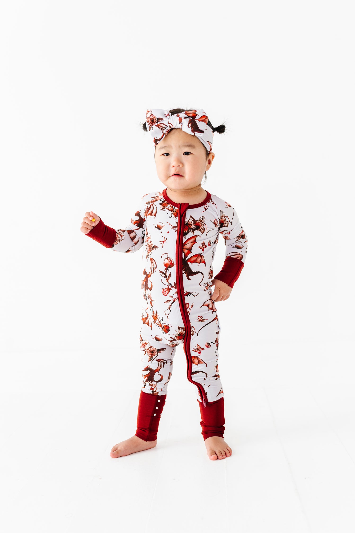 A young child stands barefoot against a white background, wearing the Jammie Session Dreaming with Dragons Romper and a matching headband.