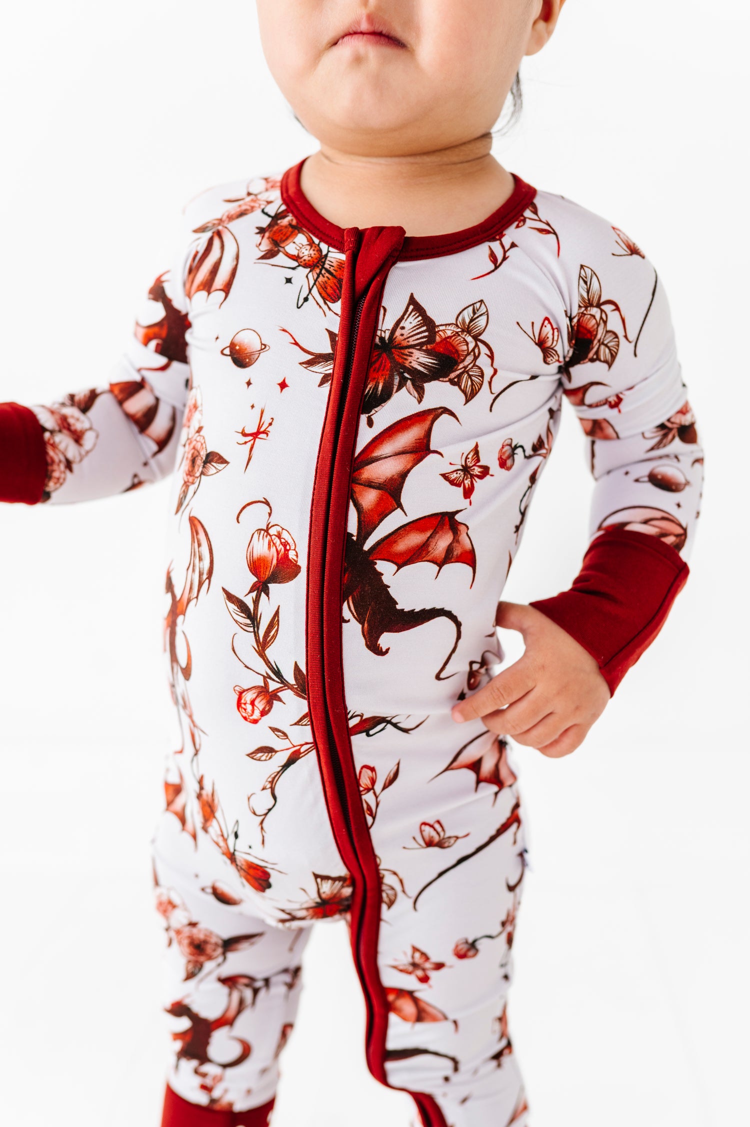 A toddler models the Jammie Session Dreaming with Dragons Romper—a white and red bamboo outfit featuring dragon and floral patterns—against a white background.