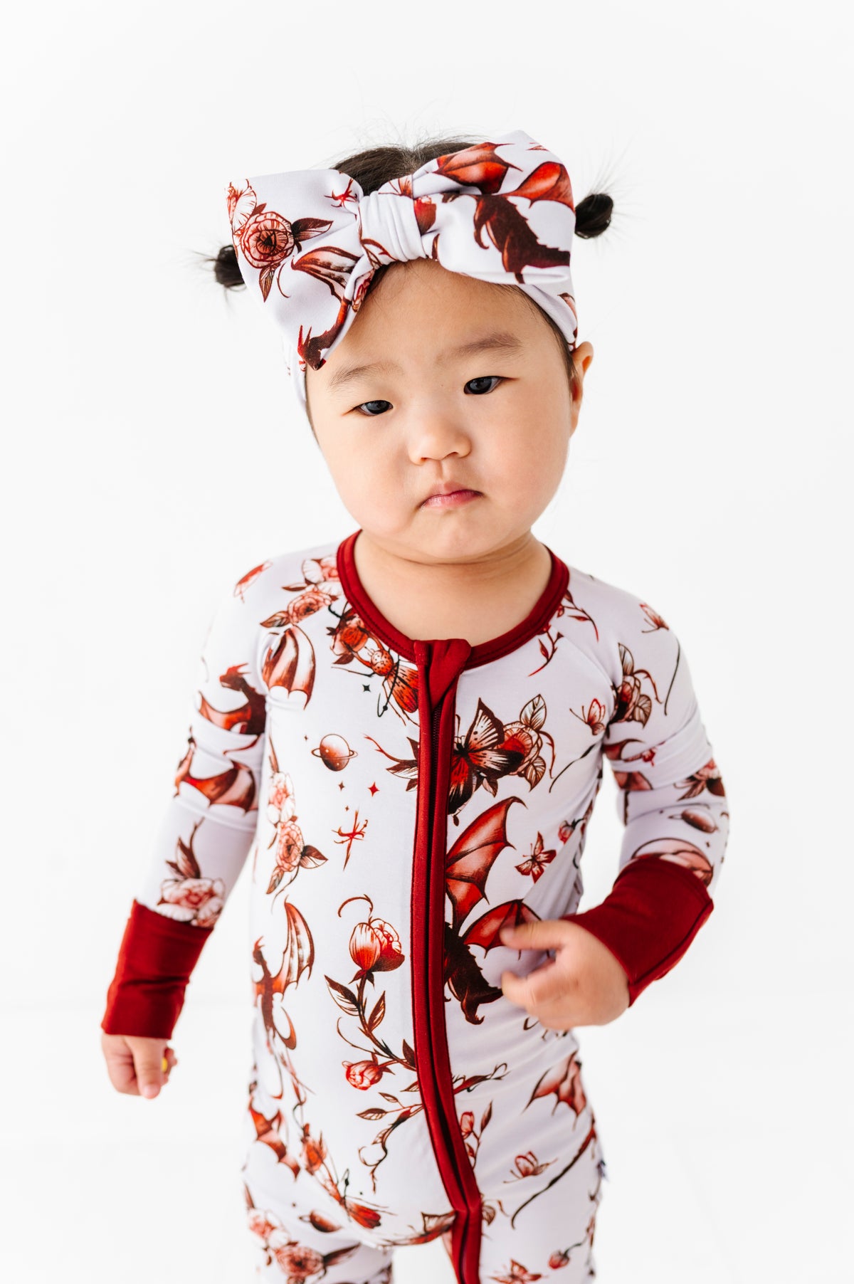 A toddler models the Jammie Session Dreaming with Dragons Romper, a white floral-patterned long sleeve outfit with red accents and matching headband, standing against a white background.