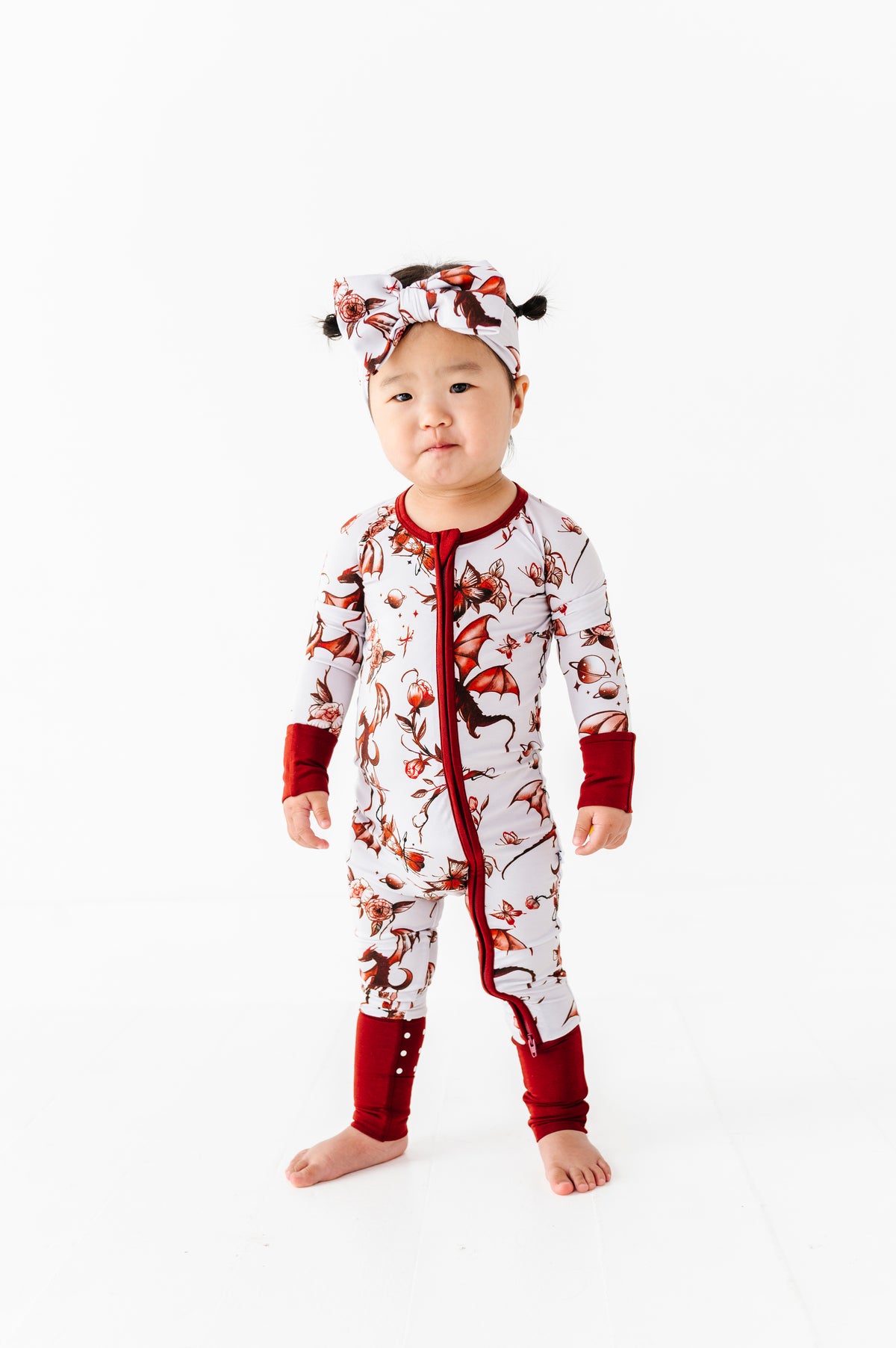 A toddler faces forward against a white background, wearing the Jammie Session "Dreaming with Dragons Romper," a red and white printed long sleeve baby romper, paired with a matching headband.