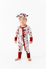 A toddler faces forward against a white background, wearing the Jammie Session "Dreaming with Dragons Romper," a red and white printed long sleeve baby romper, paired with a matching headband.