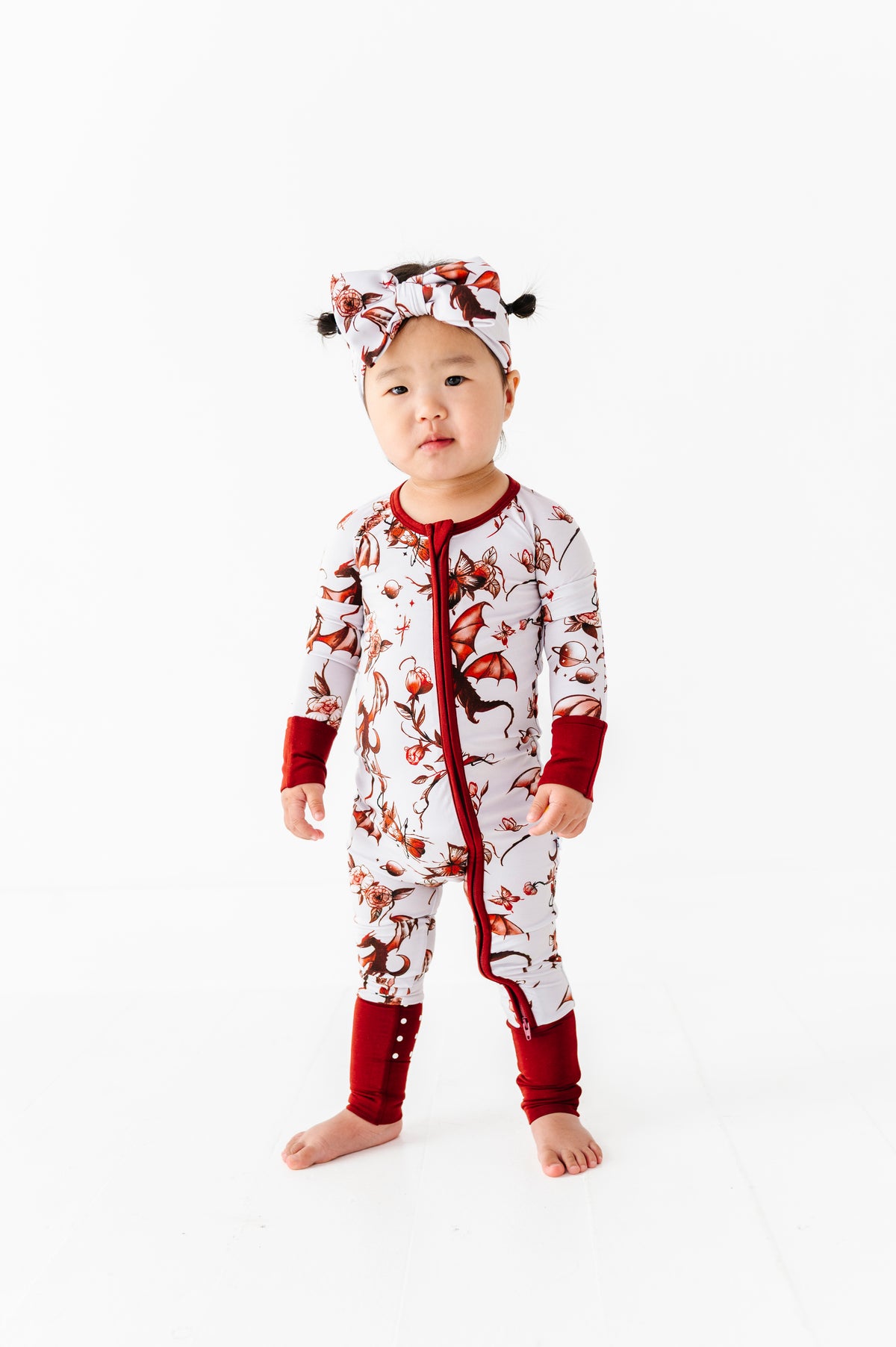A young child faces the camera in a Jammie Session "Dreaming with Dragons Romper," a white and red patterned long-sleeve baby outfit with matching headband, set against a plain white background.