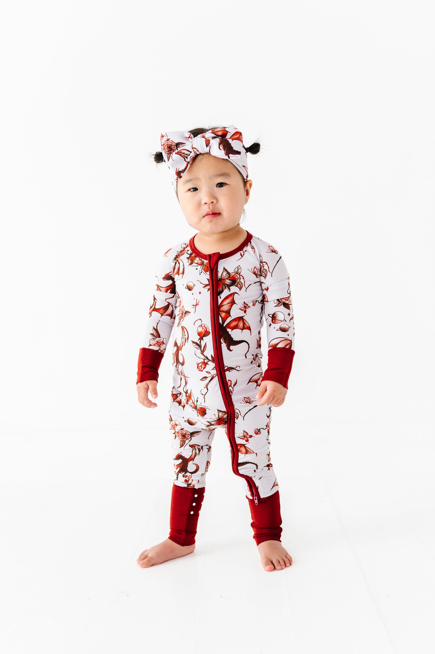 A young child faces the camera in a Jammie Session "Dreaming with Dragons Romper," a white and red patterned long-sleeve baby outfit with matching headband, set against a plain white background.