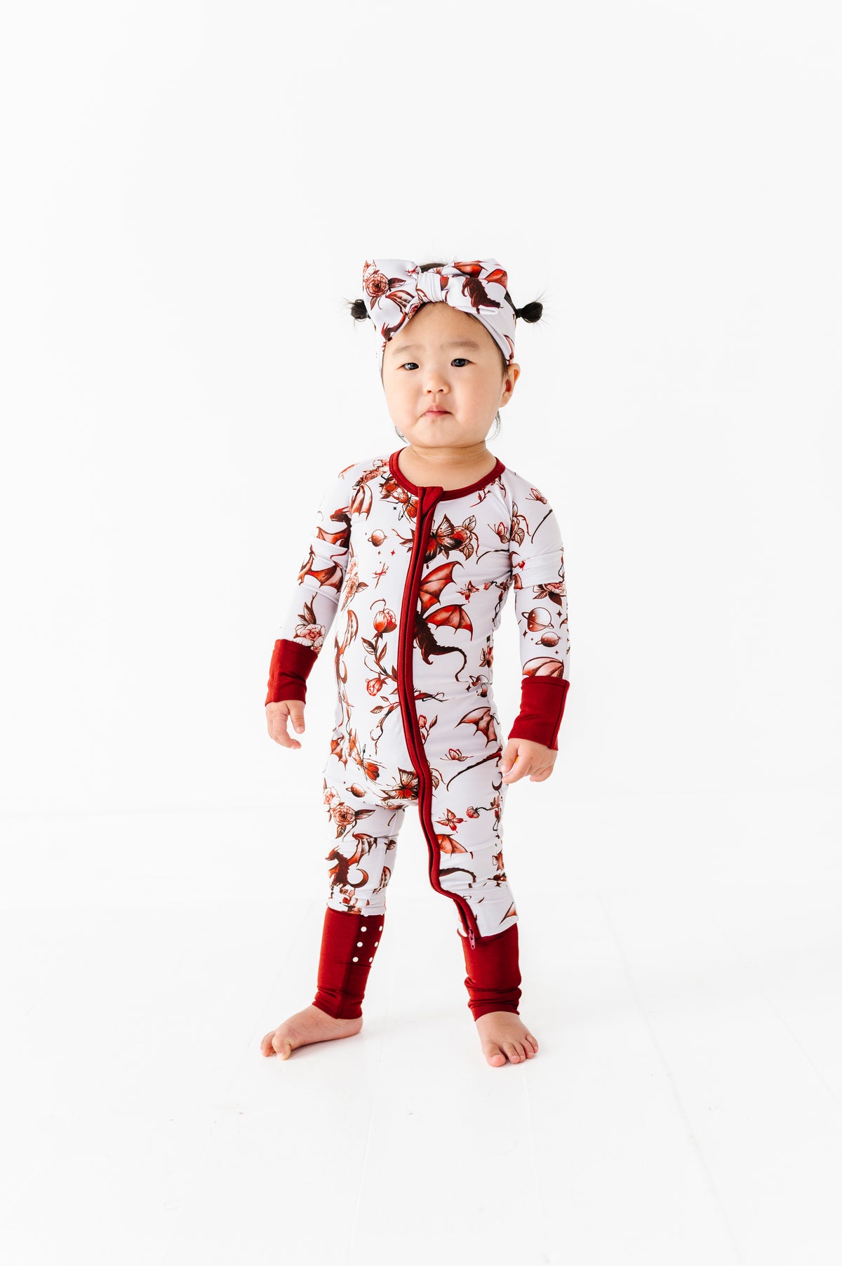 A toddler wears the Jammie Session Dreaming with Dragons Romper, a white and red bamboo pajama set with dragon print and matching headwrap, standing barefoot against a plain white background.