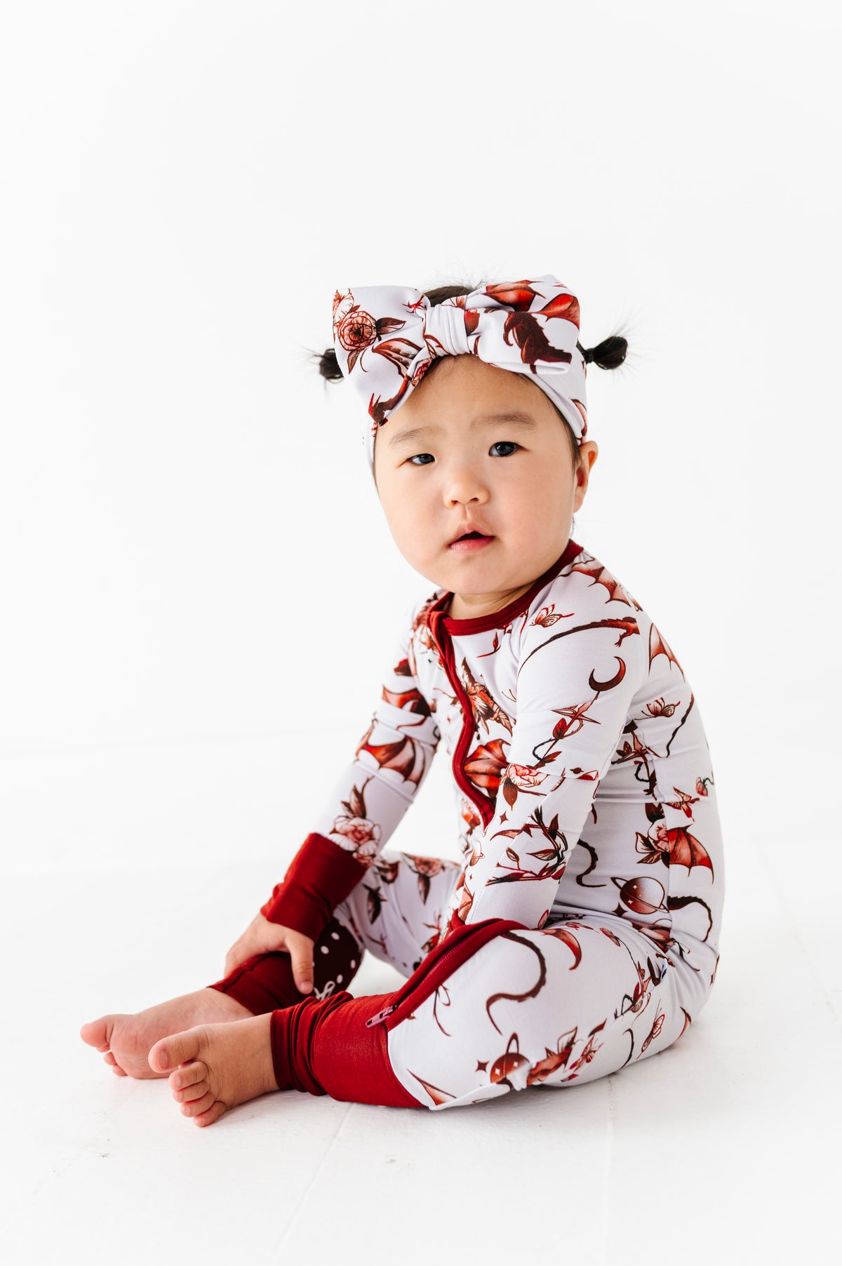 A baby wearing the Jammie Session Dreaming with Dragons Romper in a red and white floral pattern, paired with a matching headband, sits on the floor against a white background.